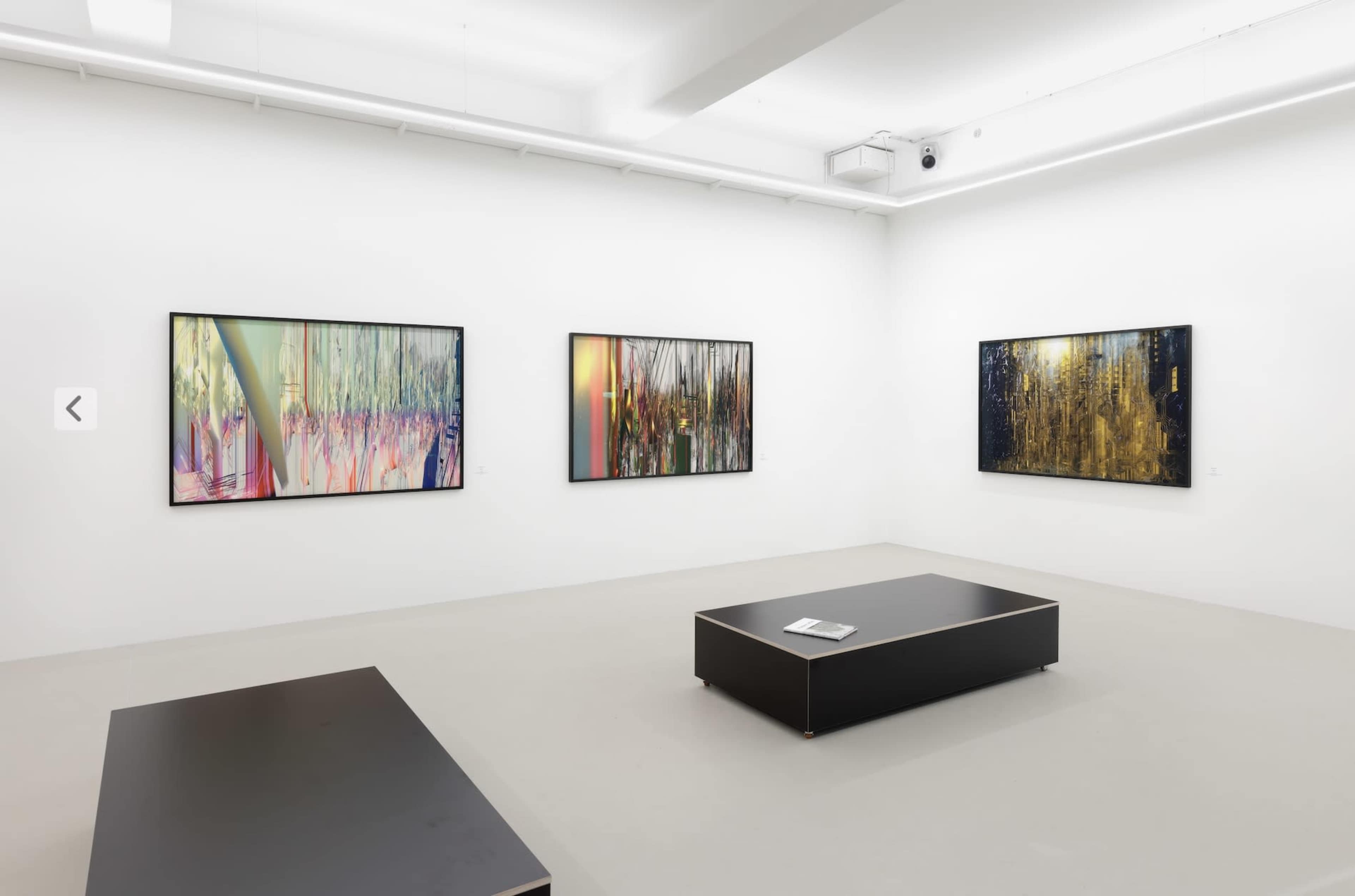 The image shows an art gallery featuring four colorful abstract paintings displayed on white walls, with a black coffee table in the center and a magazine resting on it.