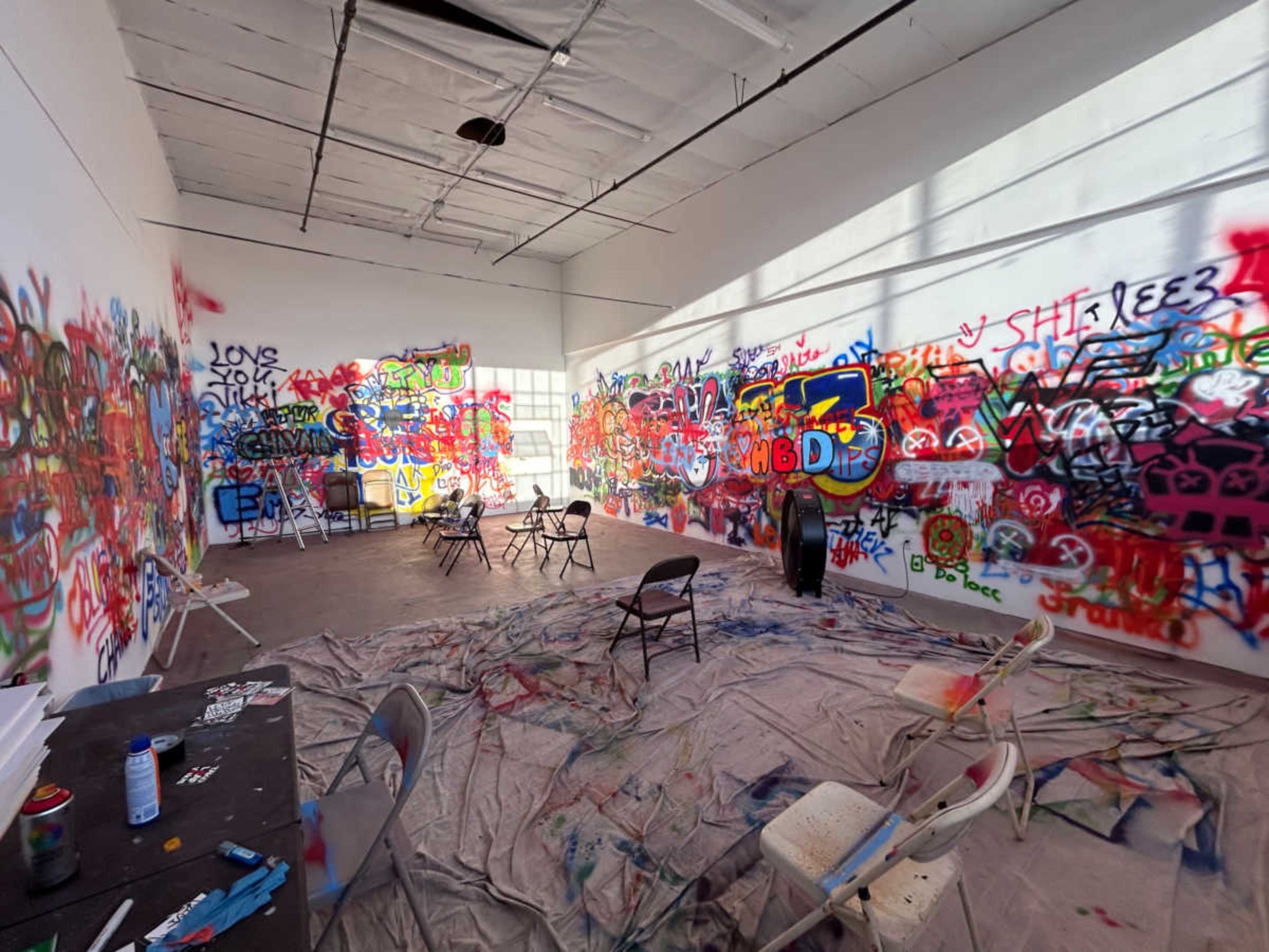 An art studio filled with colorful graffiti on white walls, chairs scattered around, and a tarp covering the floor.