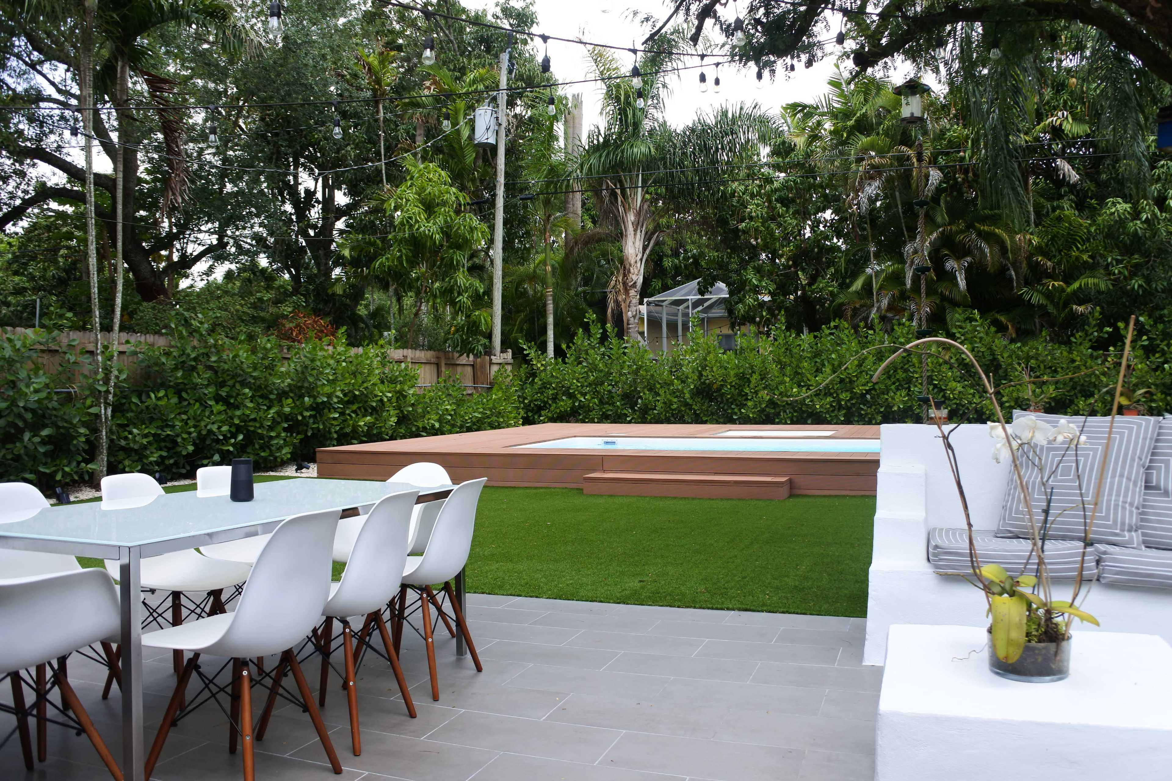 A landscaped backyard with a dining table, modern chairs, a small pool area, and lush greenery surrounding a patio space.