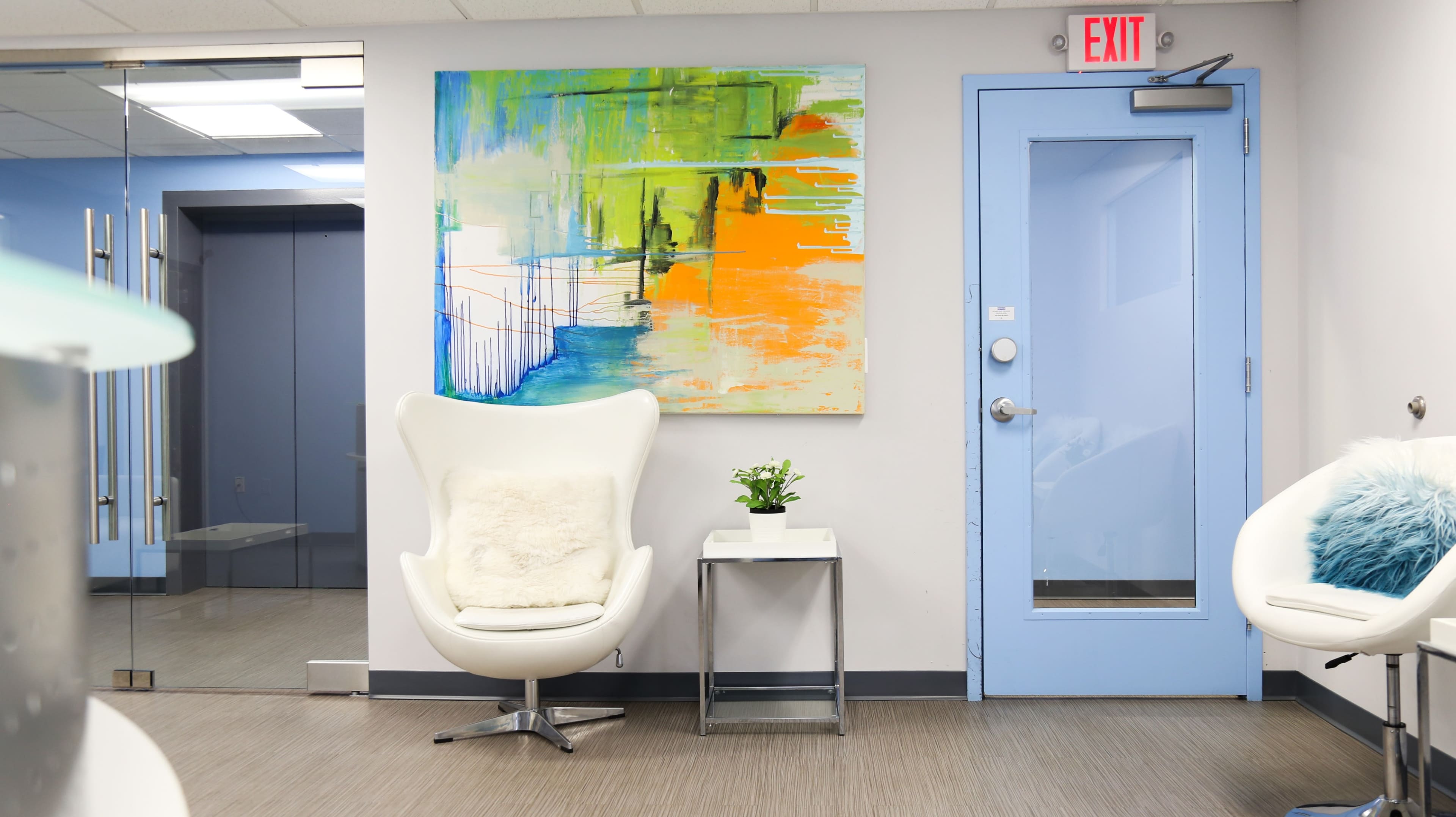 The image shows a modern waiting area featuring a colorful abstract painting, a white chair with a fur throw, a small plant on a glass table, and a blue door leading to another room.
