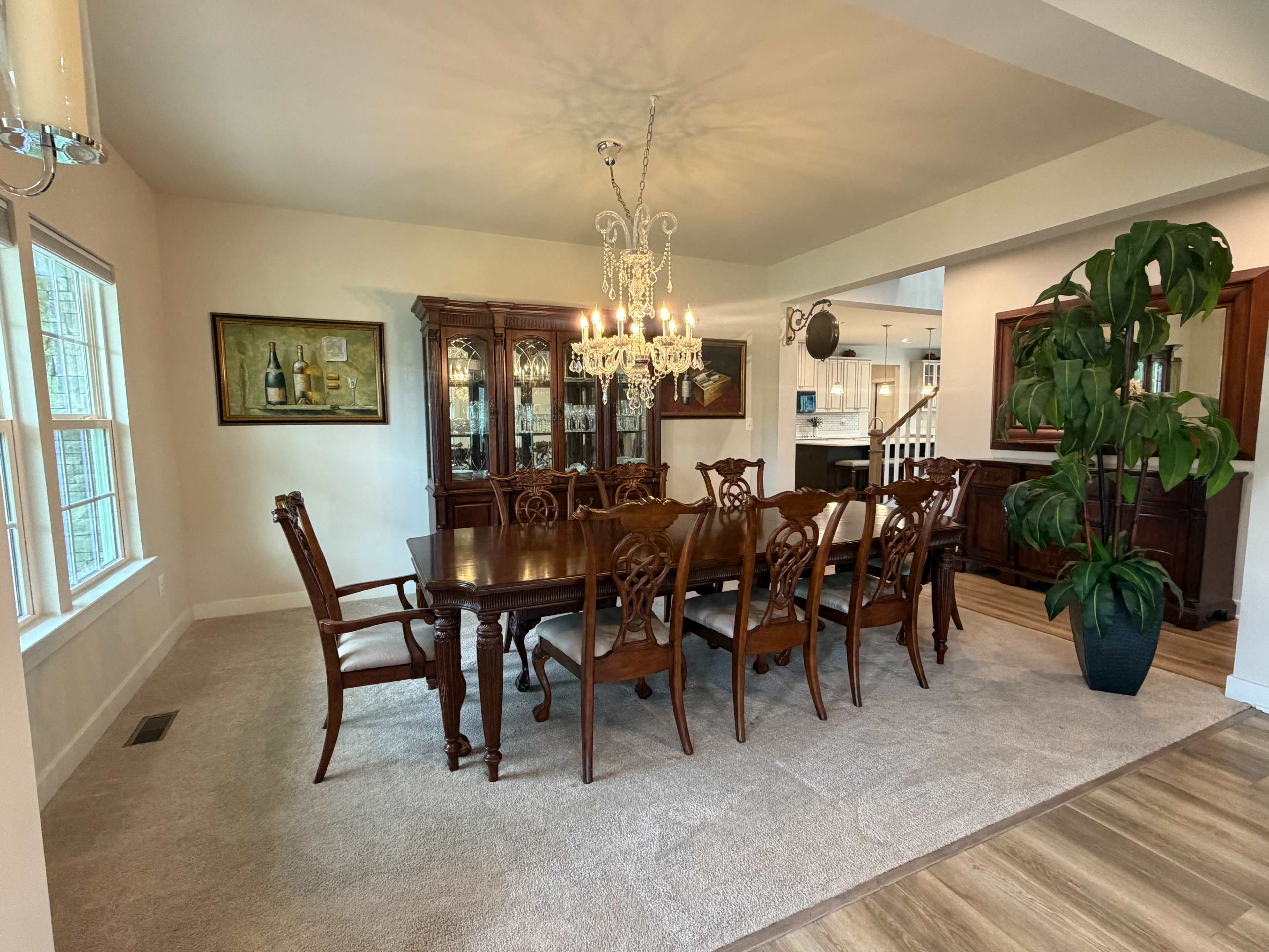 The image shows a spacious dining room featuring a large wooden table surrounded by intricately designed chairs, a chandelier overhead, and a china cabinet against one wall.