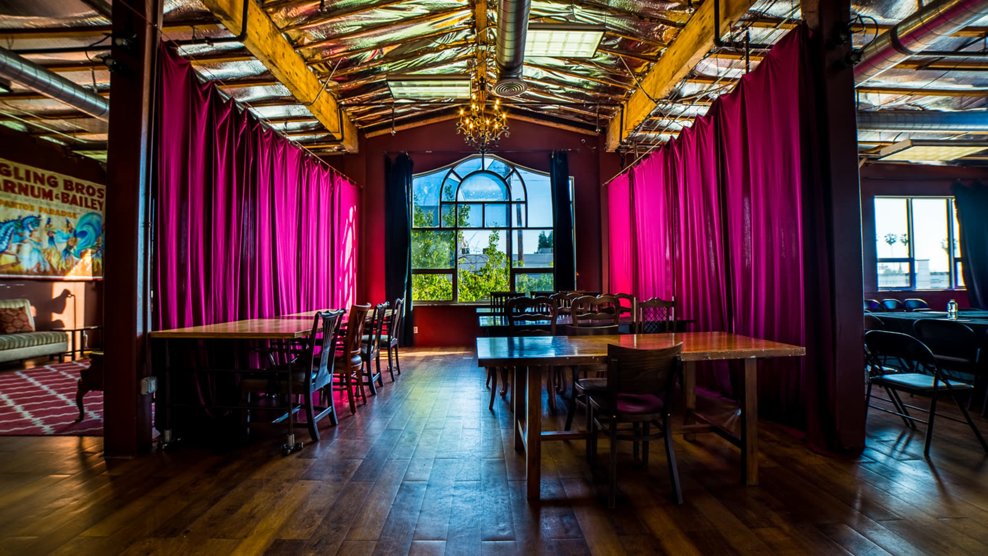 The image shows a spacious dining area with large wooden tables and vibrant pink curtains draping along the walls, illuminated by natural light from large windows.