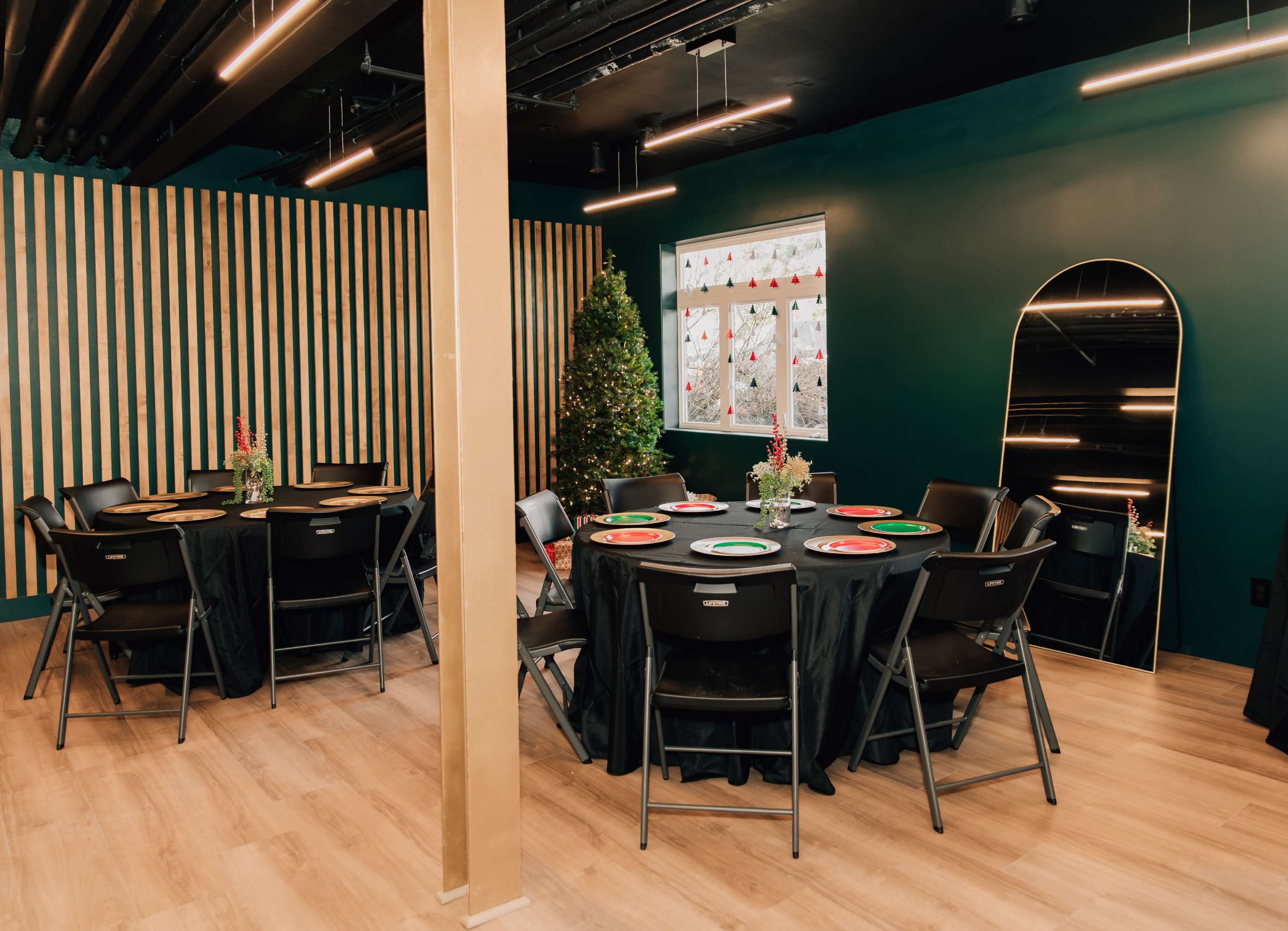The image shows a dining area with several round tables set with plates, black tablecloths, and decorative centerpieces, surrounded by green walls and wooden accents.
