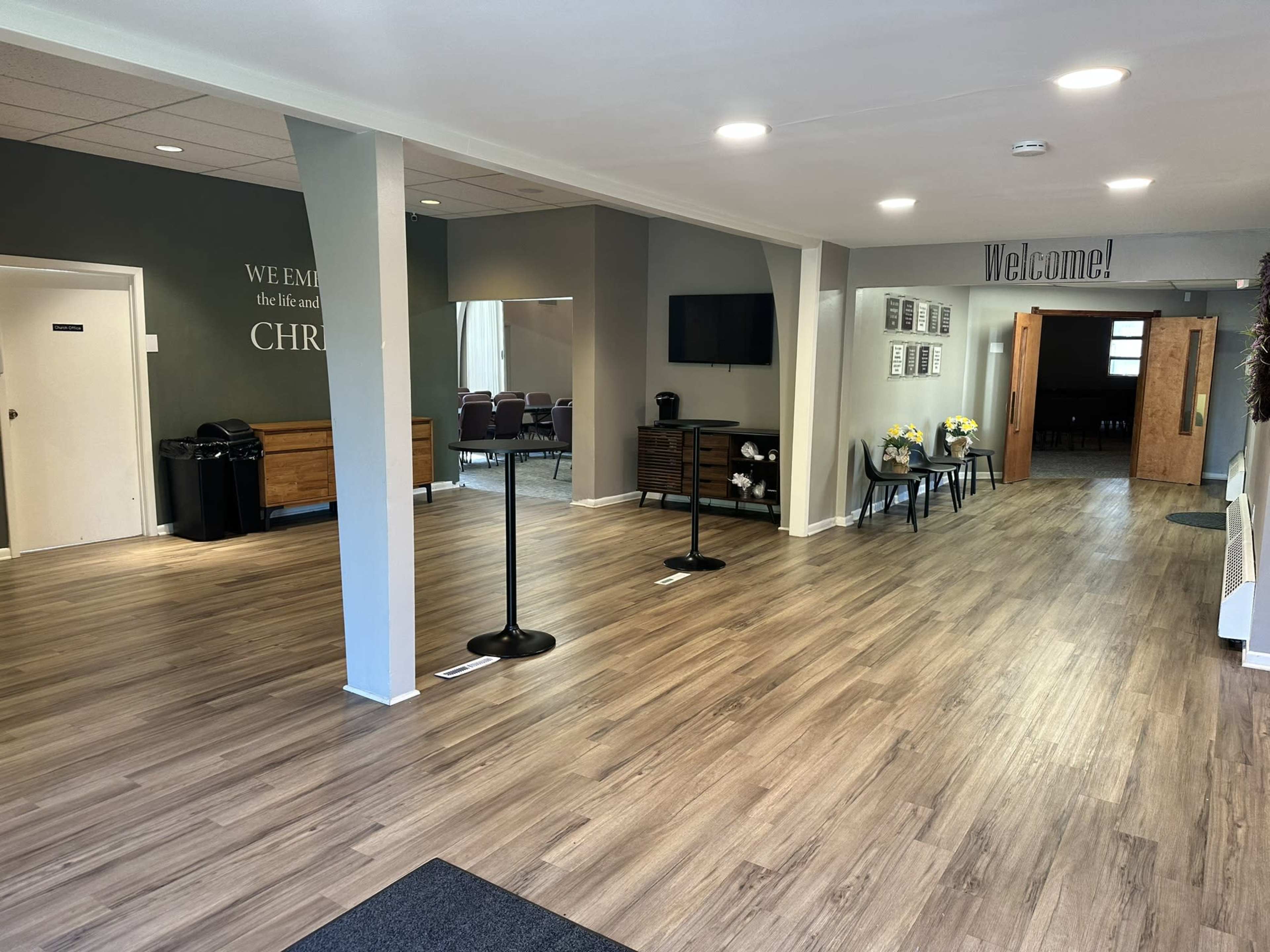 The interior of a lobby features wooden flooring, a seating area with chairs, a mirrored wall, and a welcome sign, with additional rooms visible through a doorway.