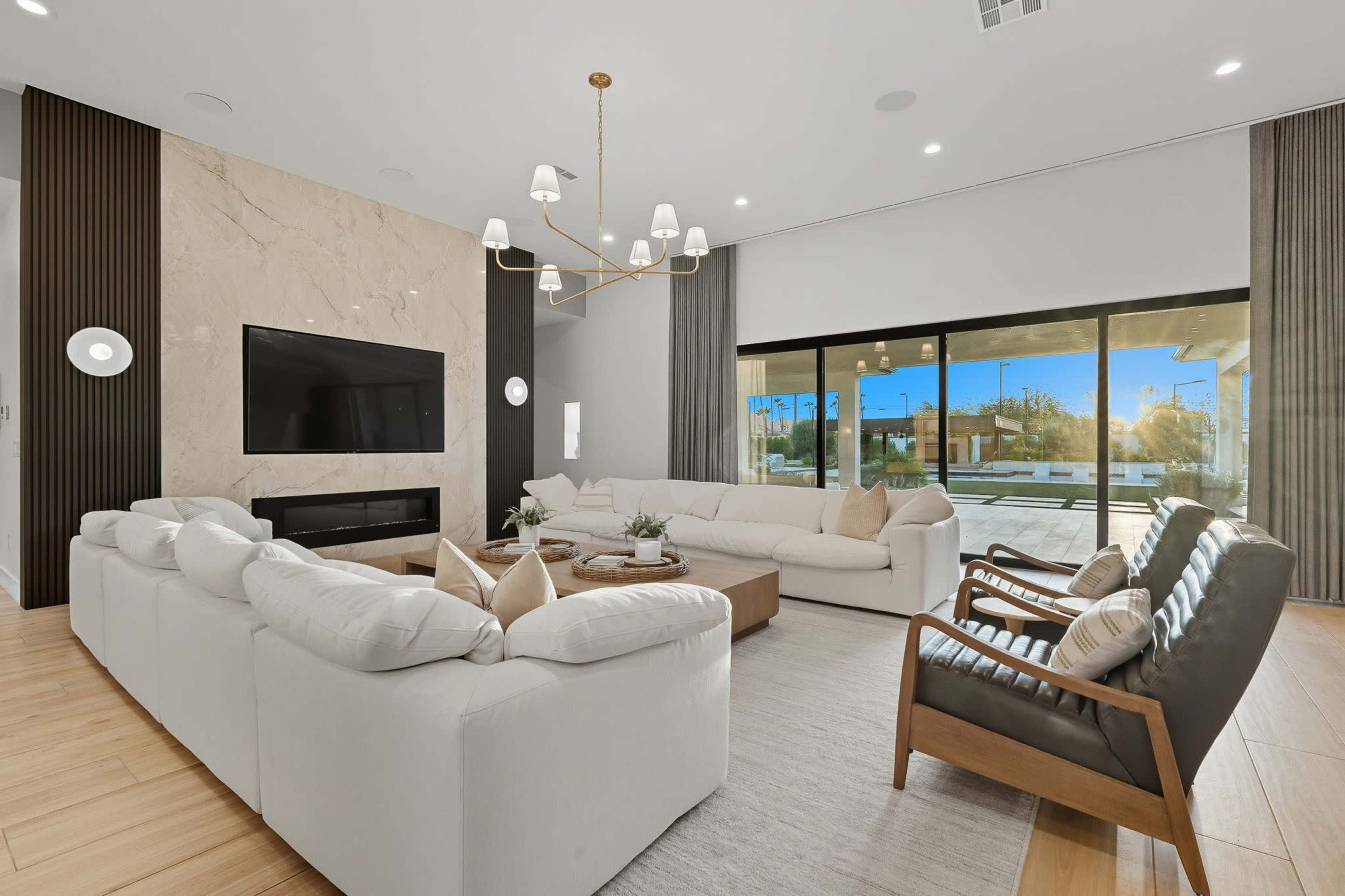 The image shows a modern living room featuring a large sectional sofa, a coffee table, and a wall-mounted television above a fireplace, with large glass doors opening to an outdoor area.