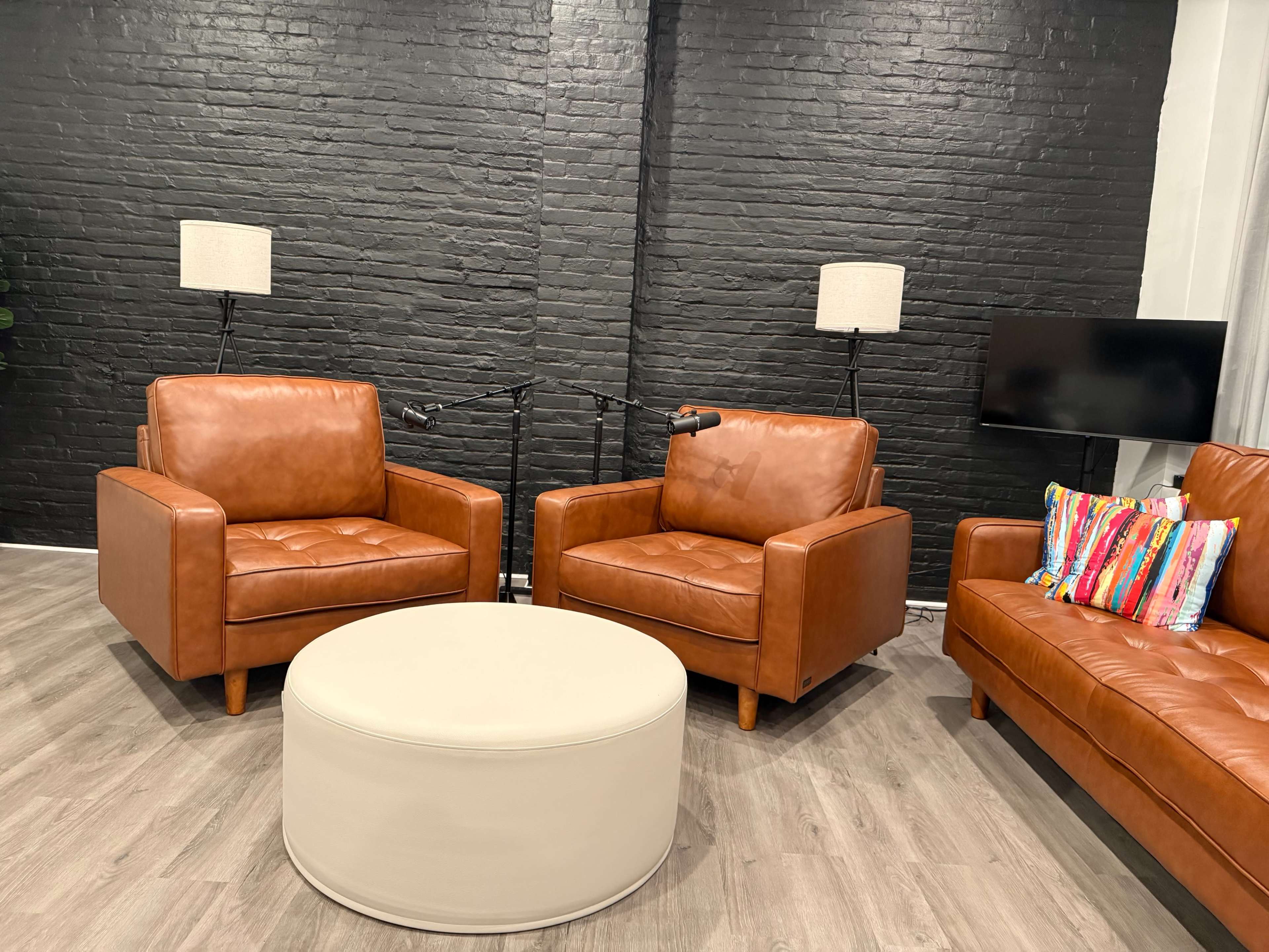 The scene shows a modern living room with two brown leather chairs, a round coffee table, and a television against a black brick wall.