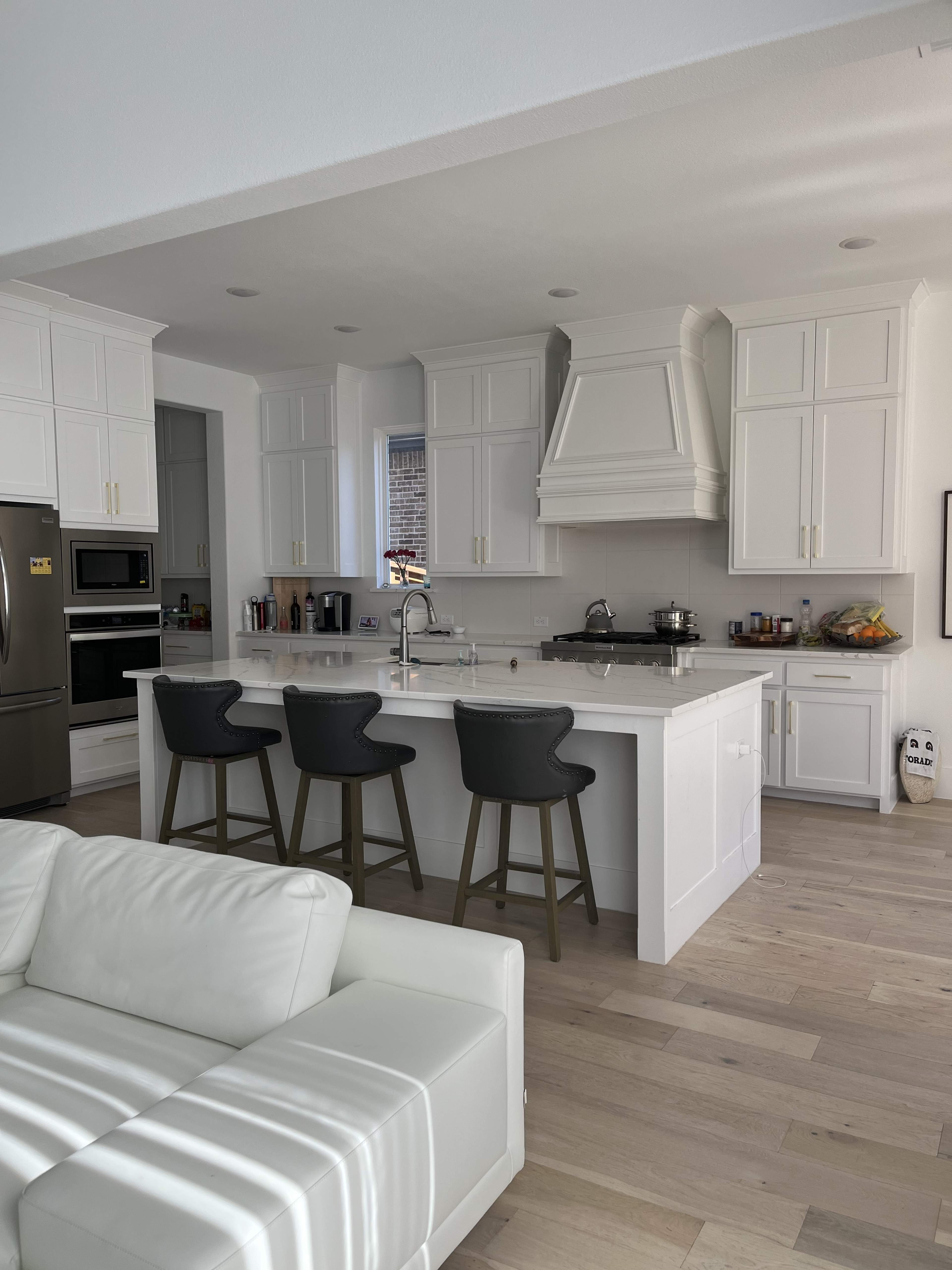 The image shows a modern kitchen with white cabinetry, a large island with four bar stools, and stainless steel appliances.