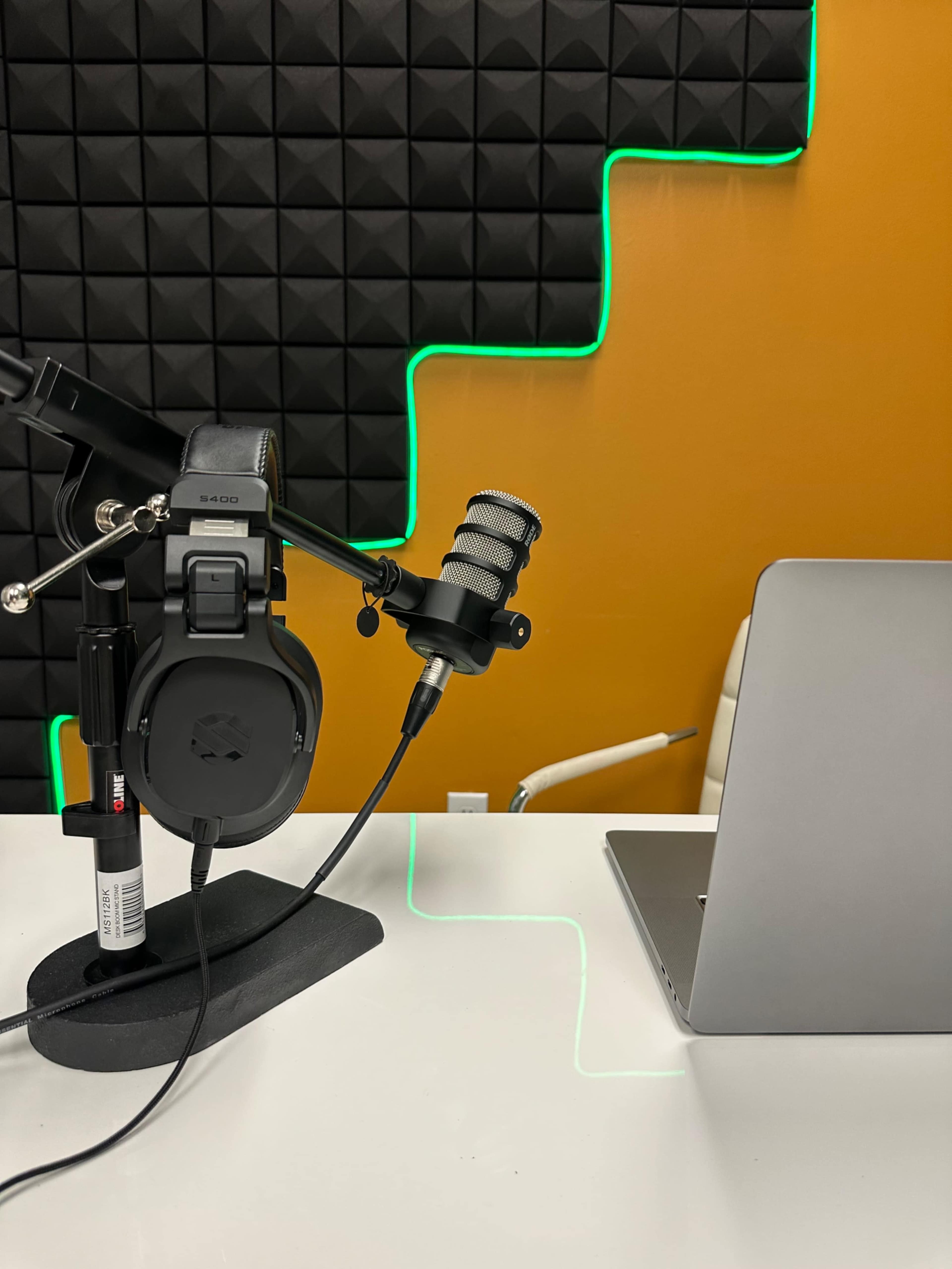 The image shows a microphone and headphones mounted on a stand next to a laptop on a desk with a textured wall and neon green lighting in the background.