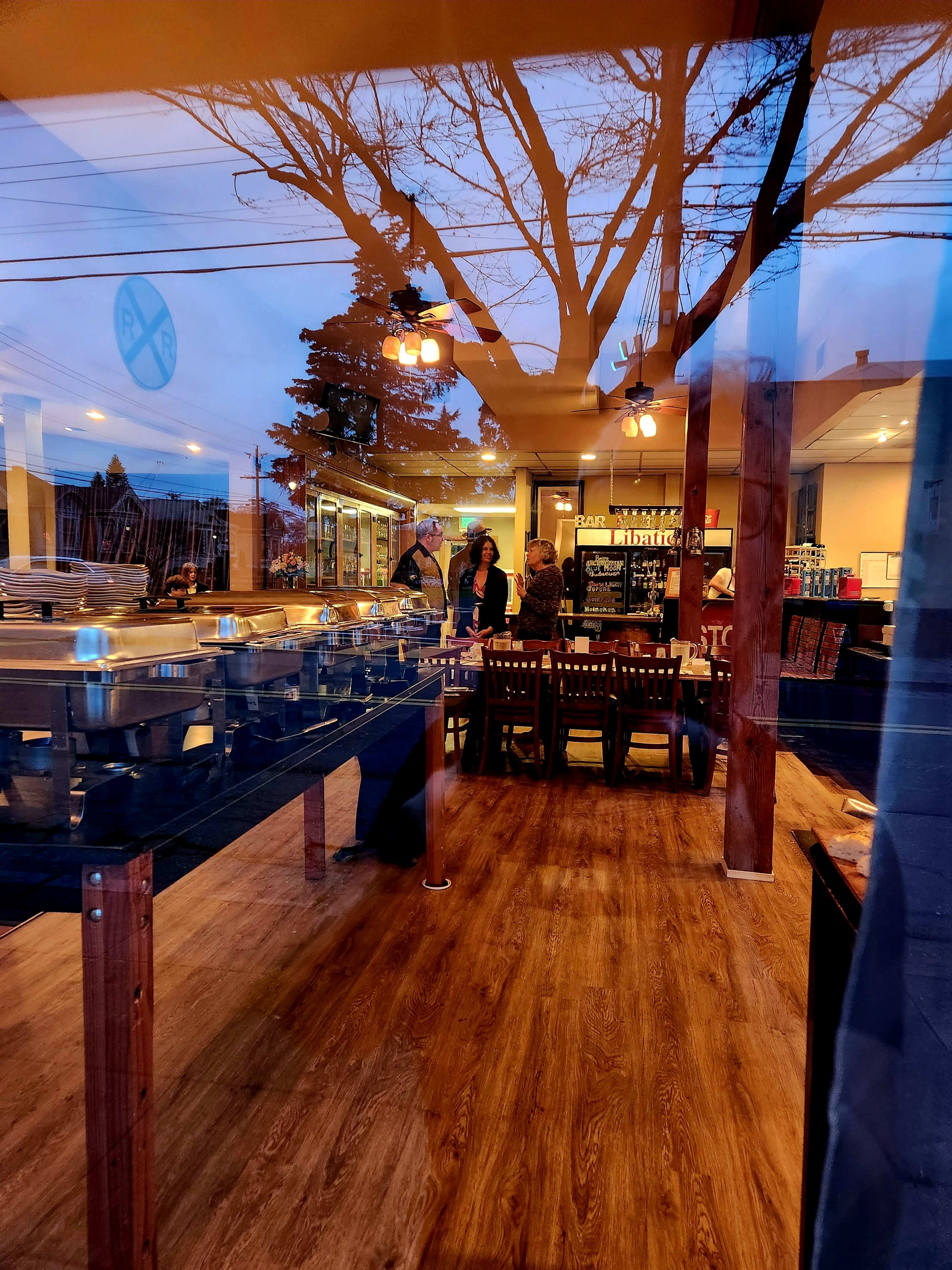 The image shows the interior of a restaurant with several buffet trays and a dining area illuminated by warm lights, viewed through reflective glass.