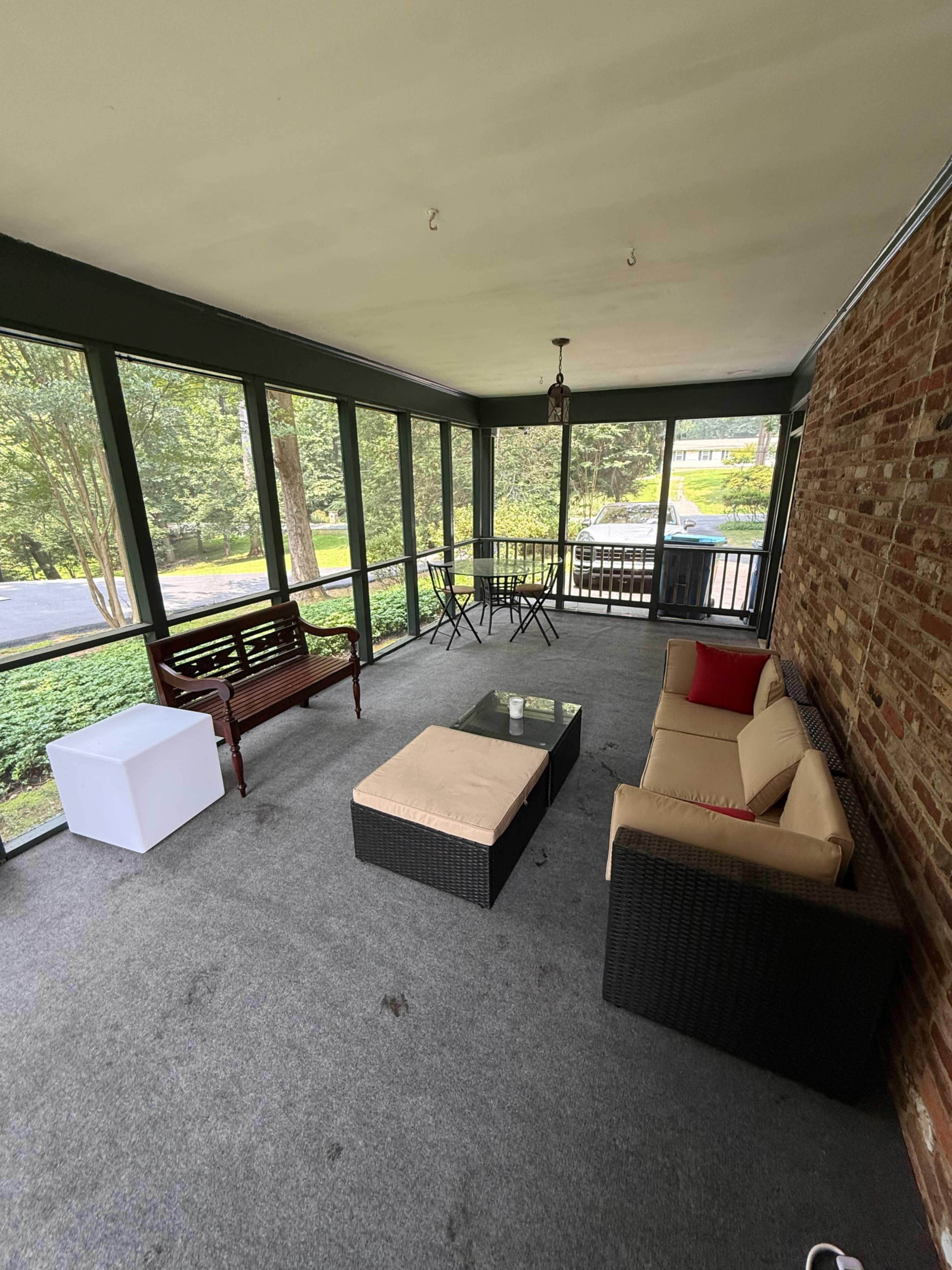 The image shows a screened-in porch with a seating area that includes a sofa, a coffee table, and a dining set, surrounded by greenery visible through the screens.