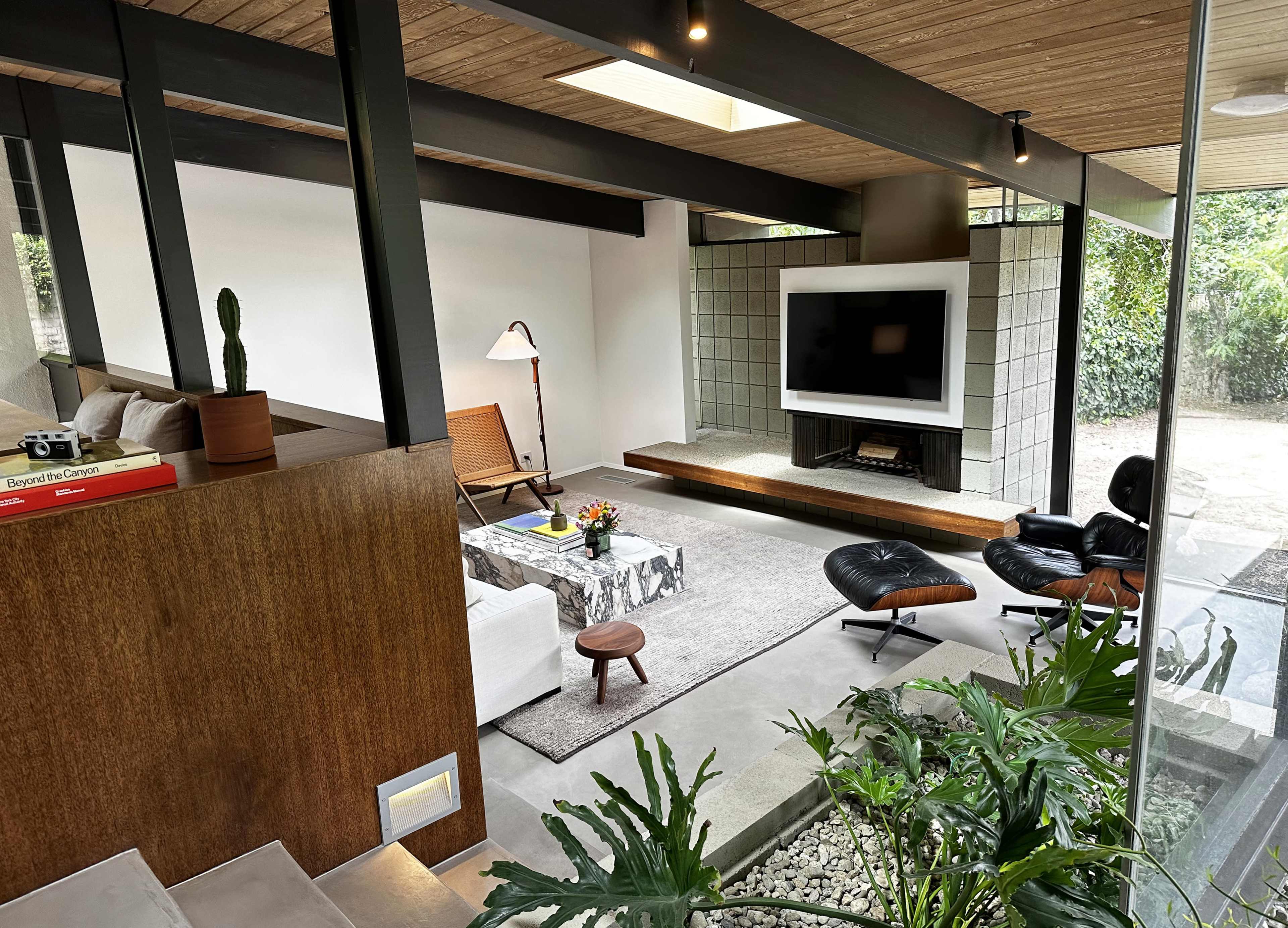 A modern living room features a fireplace, a large television, minimalist furniture, and indoor plants, with natural light streaming through large windows.