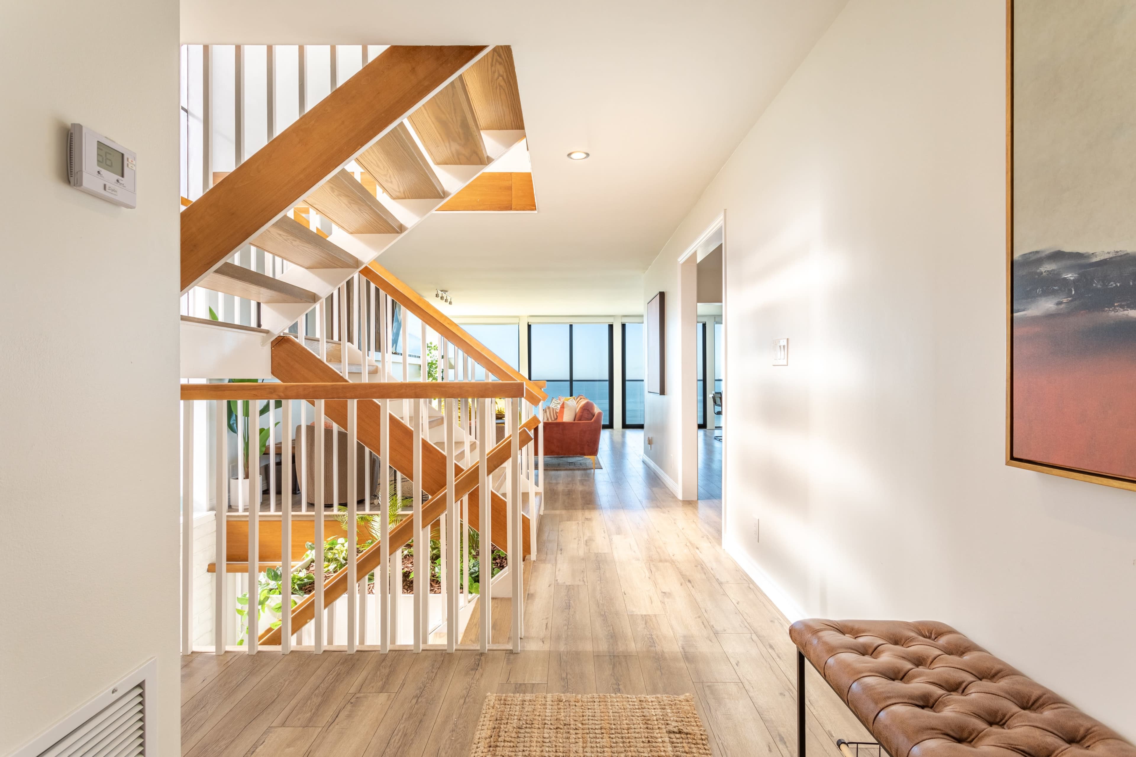 A hallway featuring a wooden staircase, a seating area in the distance, and natural light illuminating the space.