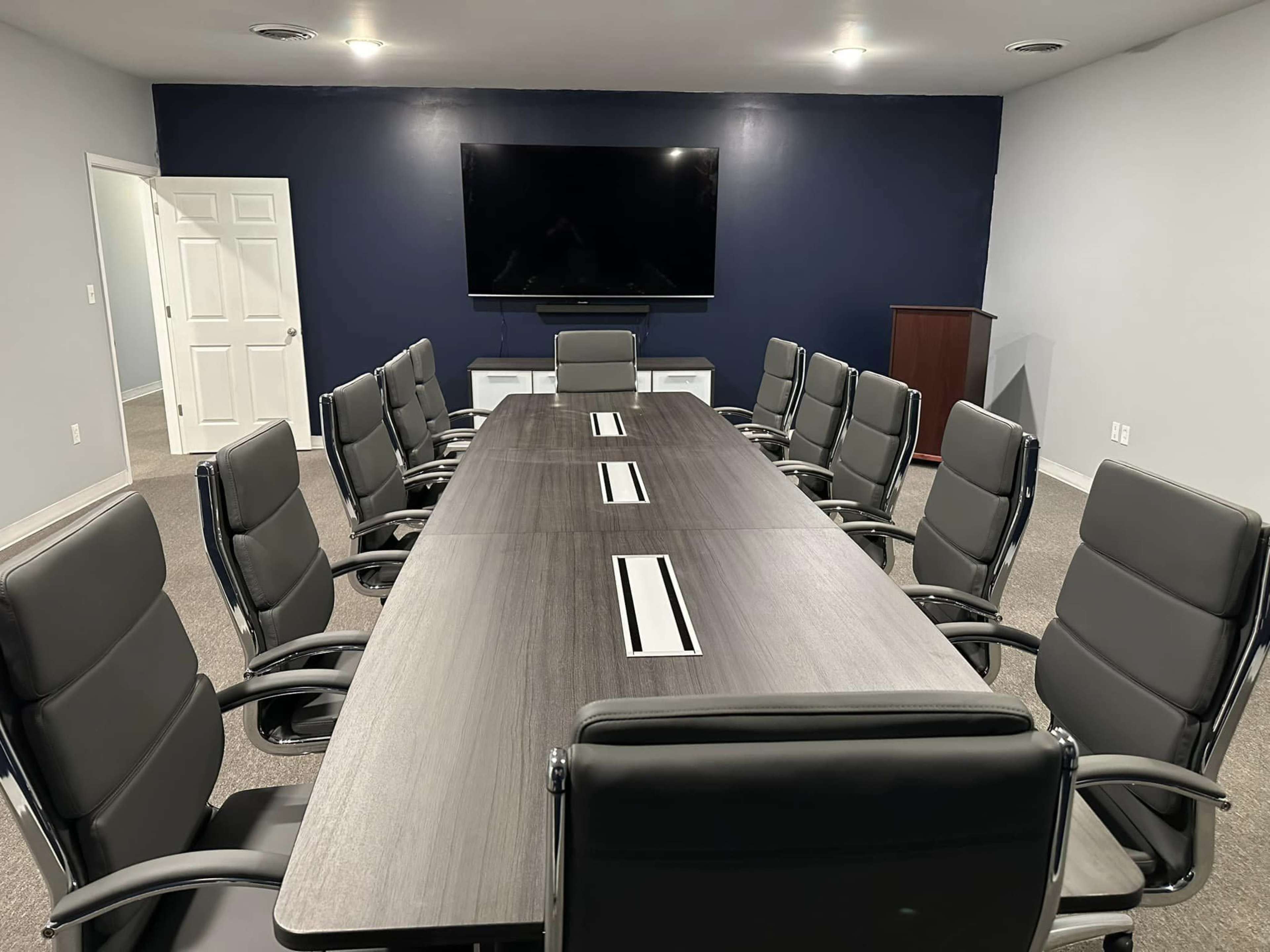 A conference room with a large rectangular table surrounded by eight chairs, a wall-mounted television, and a dark blue accent wall.