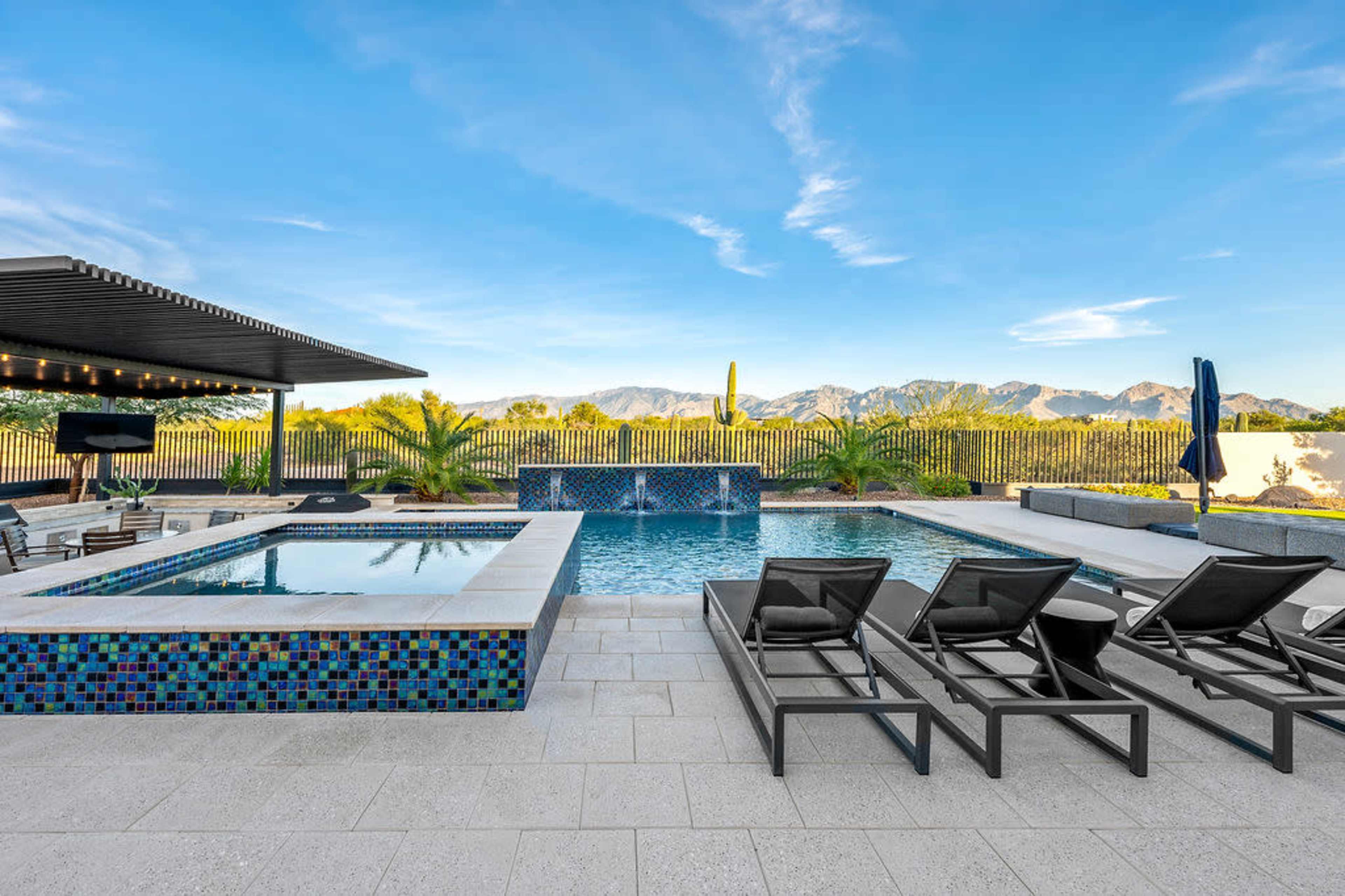 The image shows a modern outdoor pool area with lounge chairs, a spa section, and a backdrop of mountains under a clear blue sky.