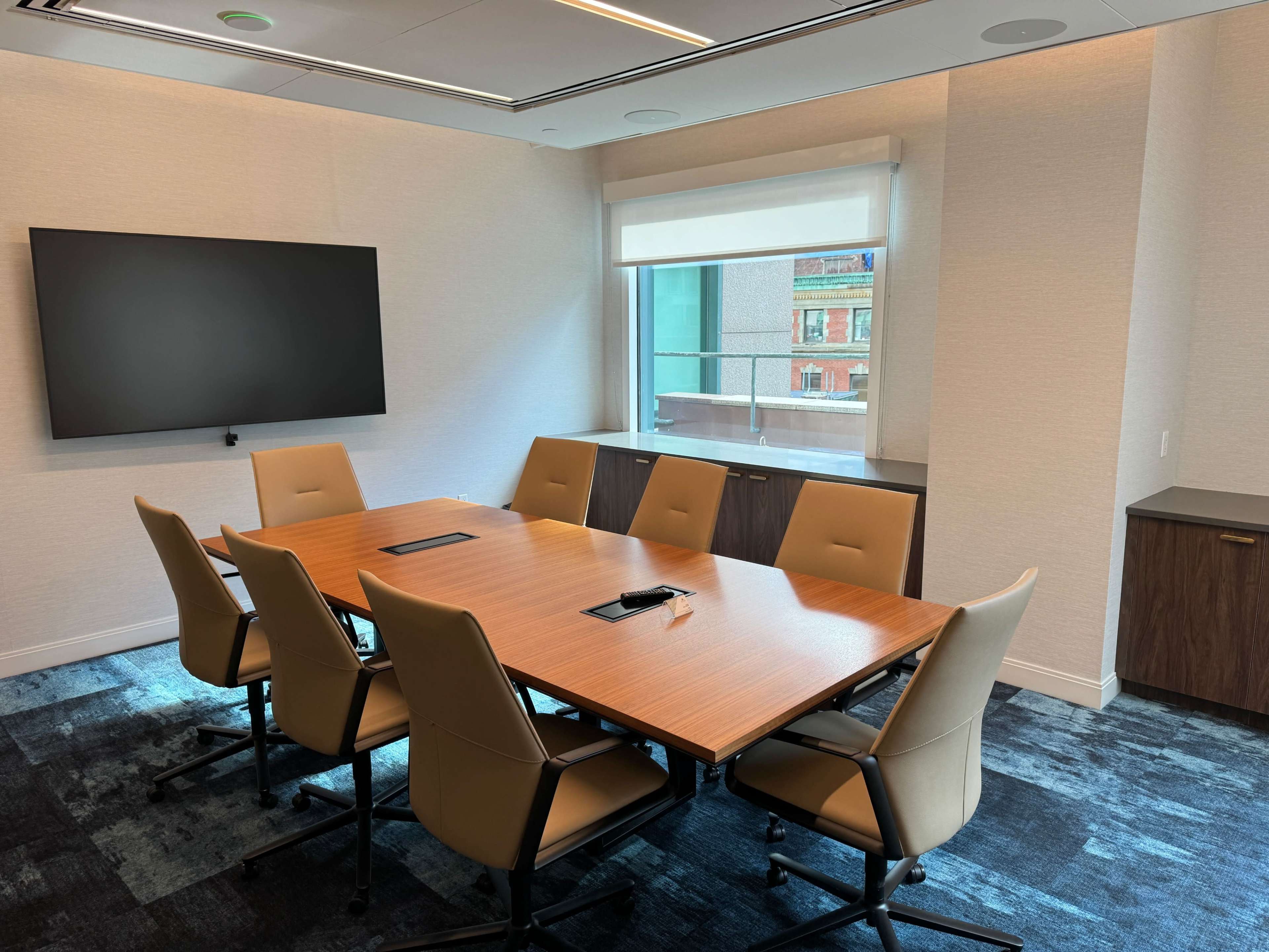 A modern conference room features a wooden table surrounded by eight light-colored chairs, with a large screen on the wall and a window overlooking a cityscape.