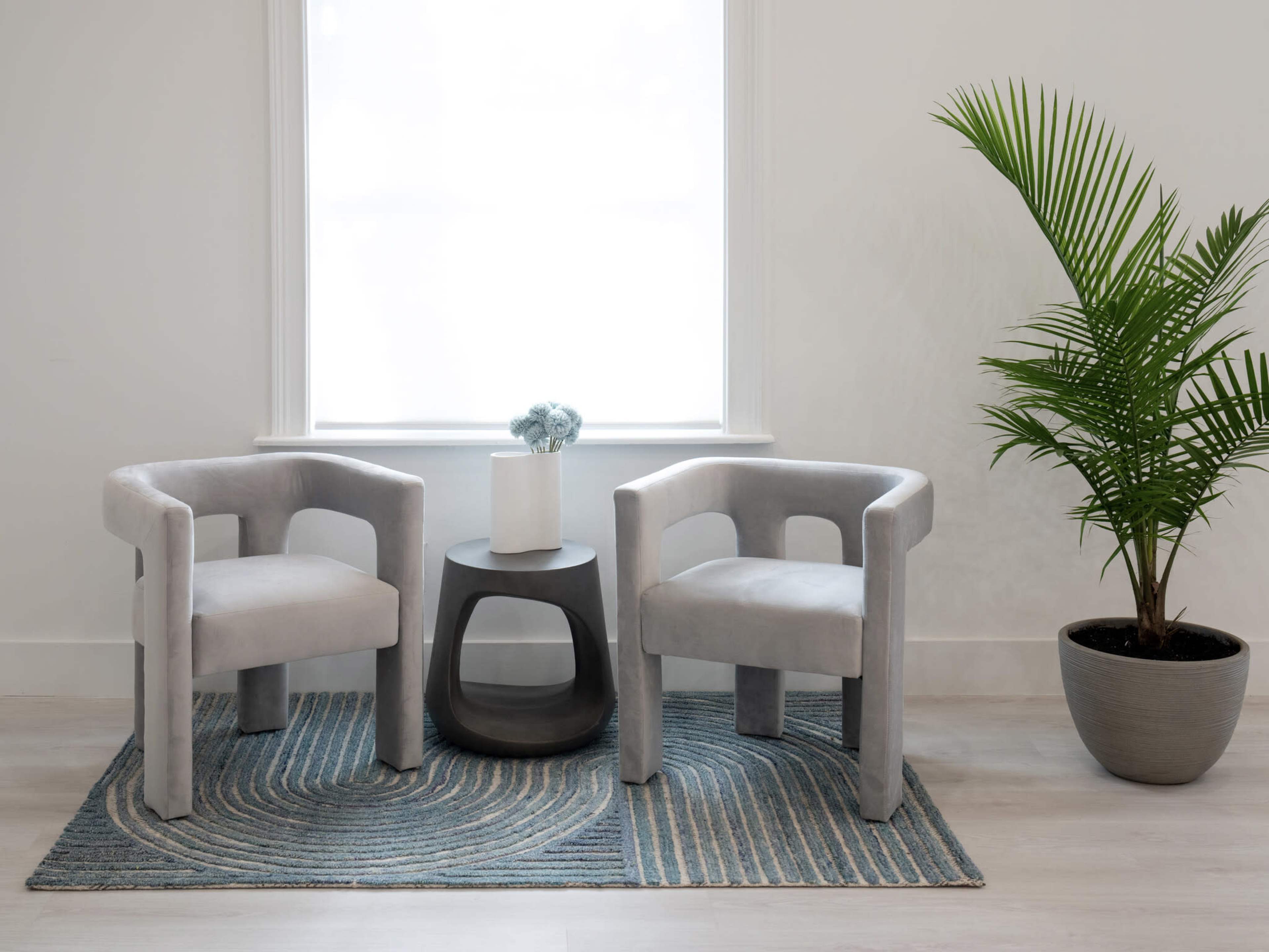 Two gray armchairs facing each other across a small black table, situated on a patterned rug near a window with a plant in a pot beside them.
