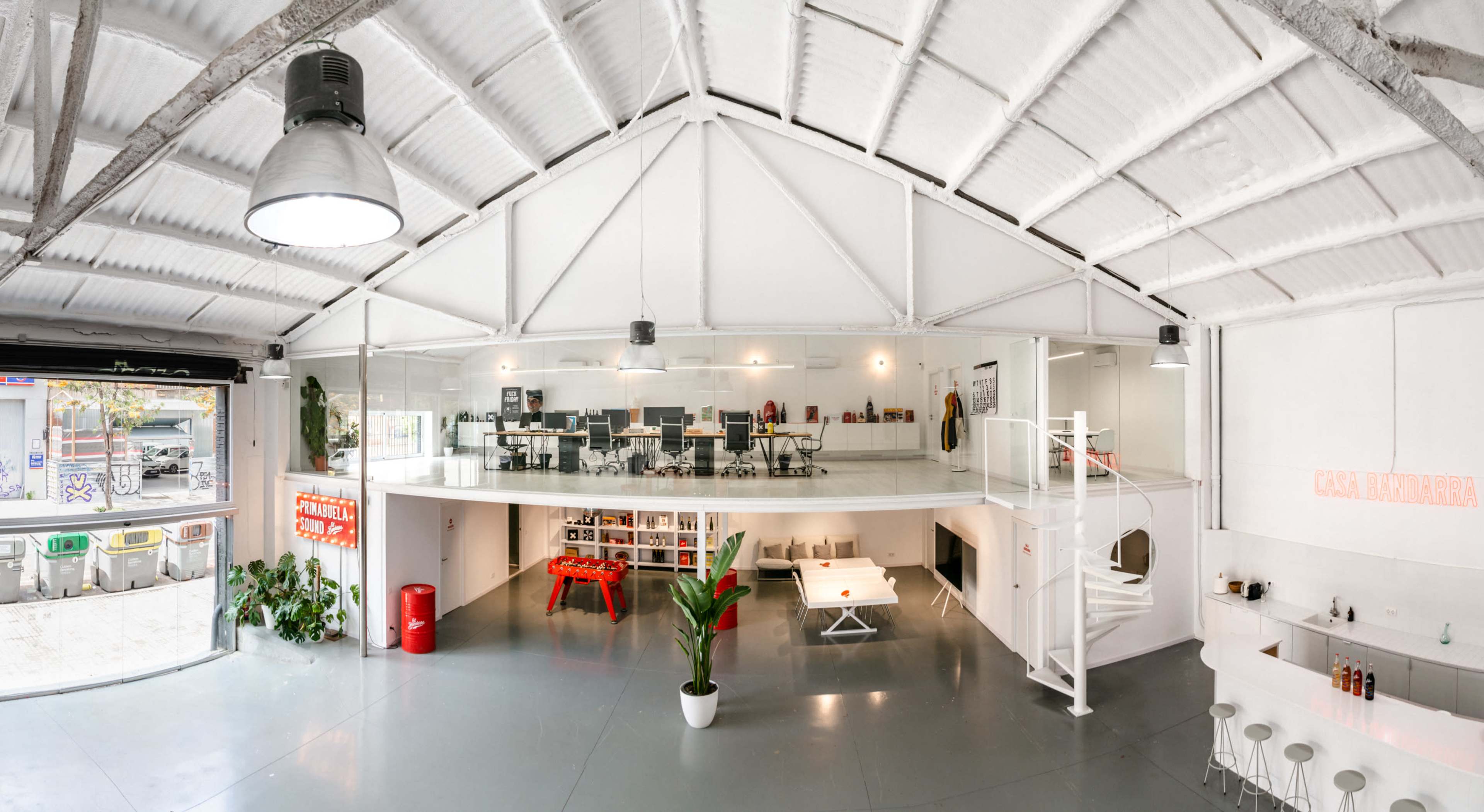 A modern open office space with a high ceiling, featuring a loft area above workstations and a communal area, along with a bar and seating arrangement on the lower level.