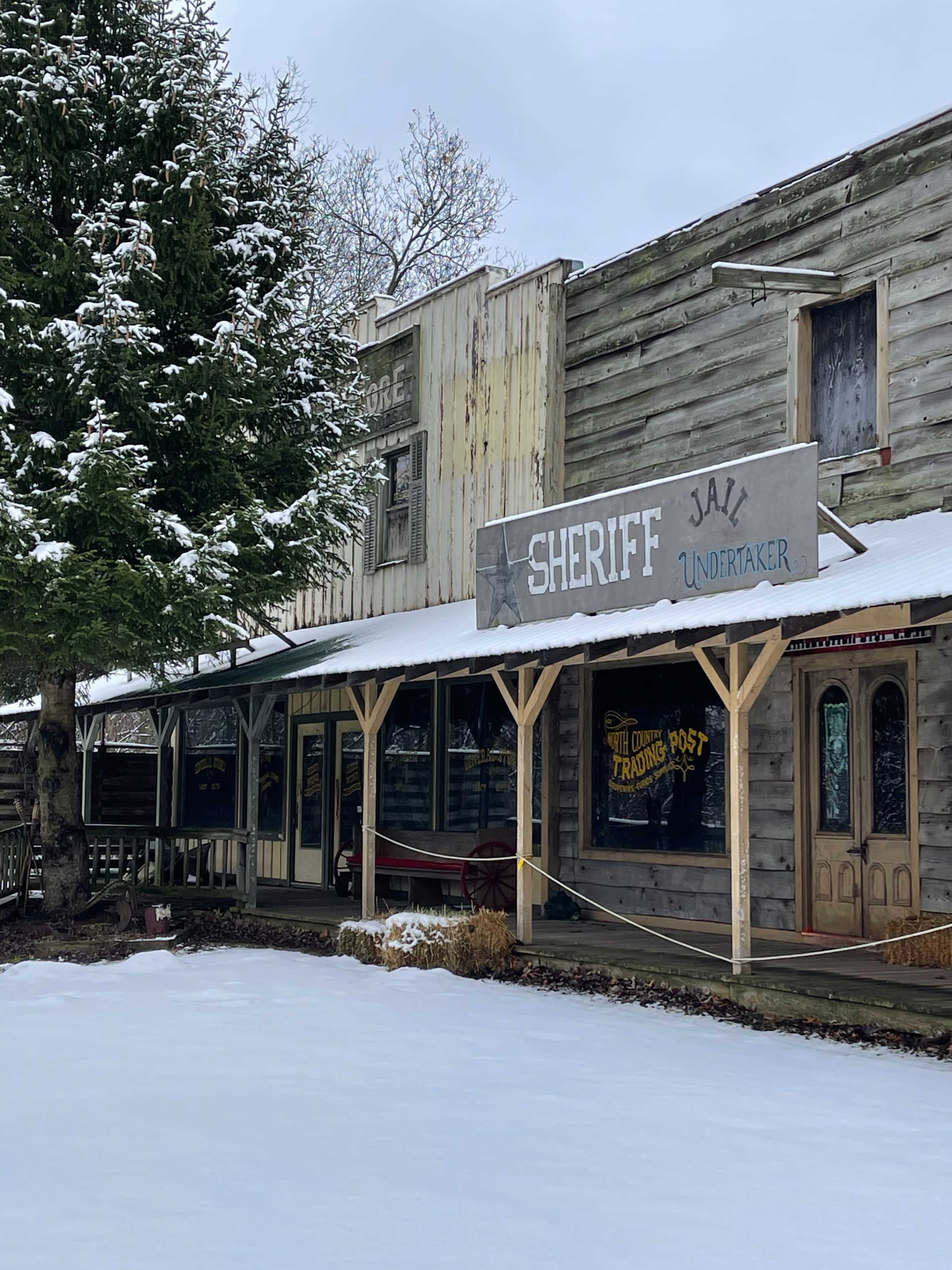 A historic wooden storefront, featuring a sign for "Sheriff Jail Undertaker," is surrounded by snow and bare trees.
