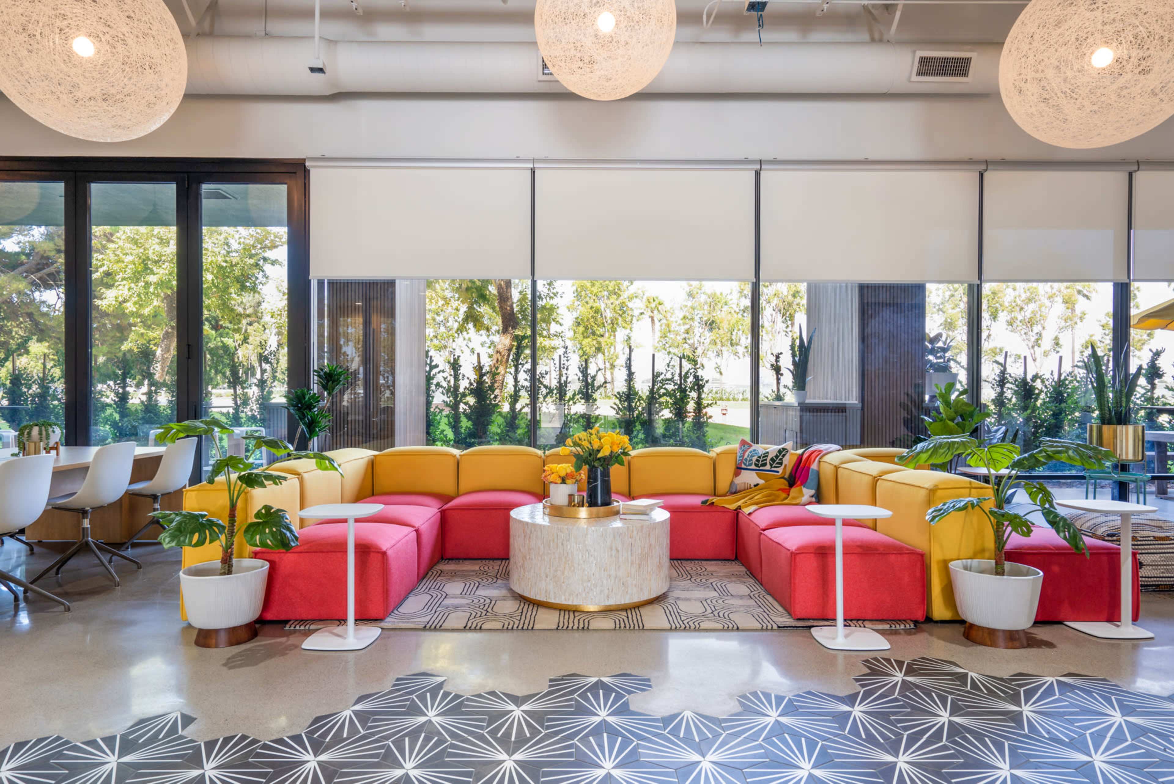 The image shows a colorful lounge area featuring a yellow and red sectional sofa, a round coffee table, and potted plants, with large windows providing a view of greenery outside.
