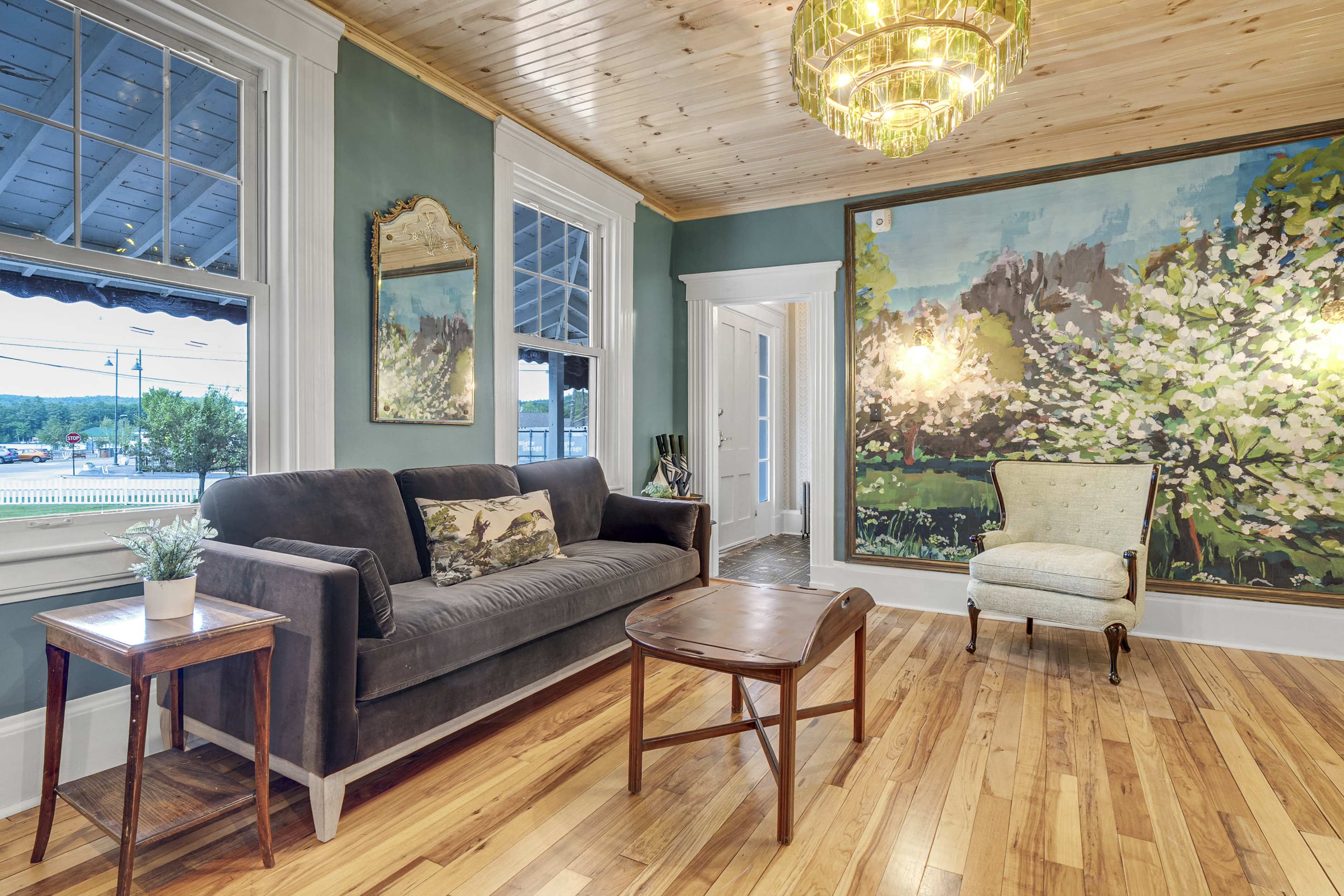 A living room features a dark couch, a small wooden table, and a light-colored chair against a backdrop of a large, vibrant landscape painting.