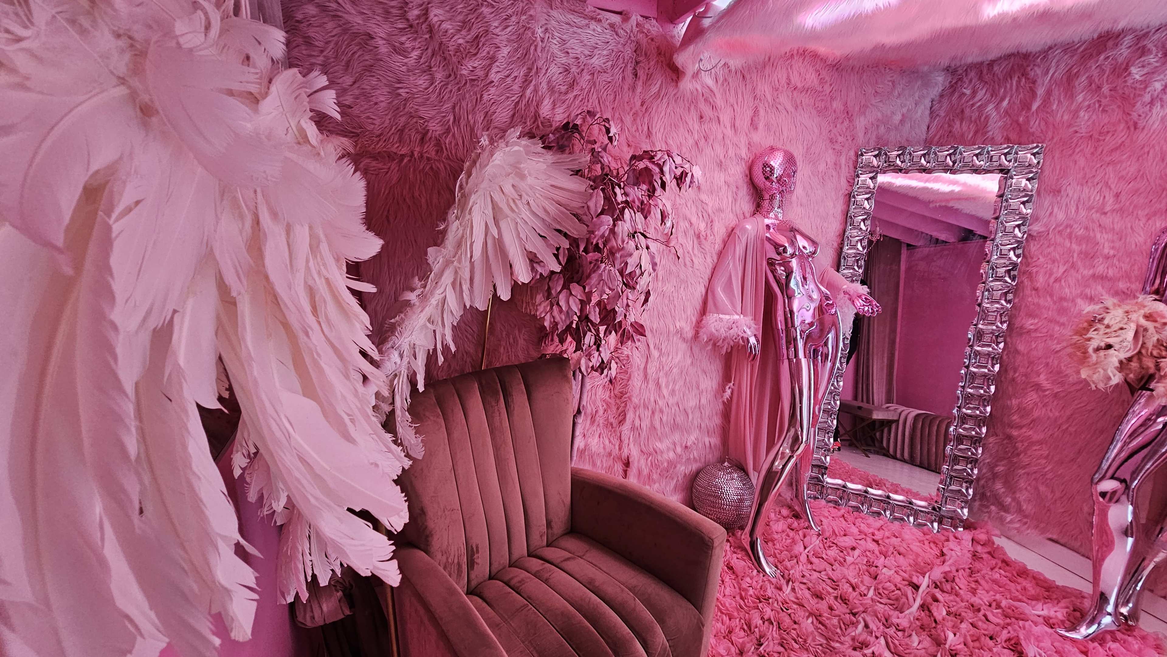 The scene features a pink-themed room with plush walls, a vintage chair, a full-length mirror, and decorative feathers.