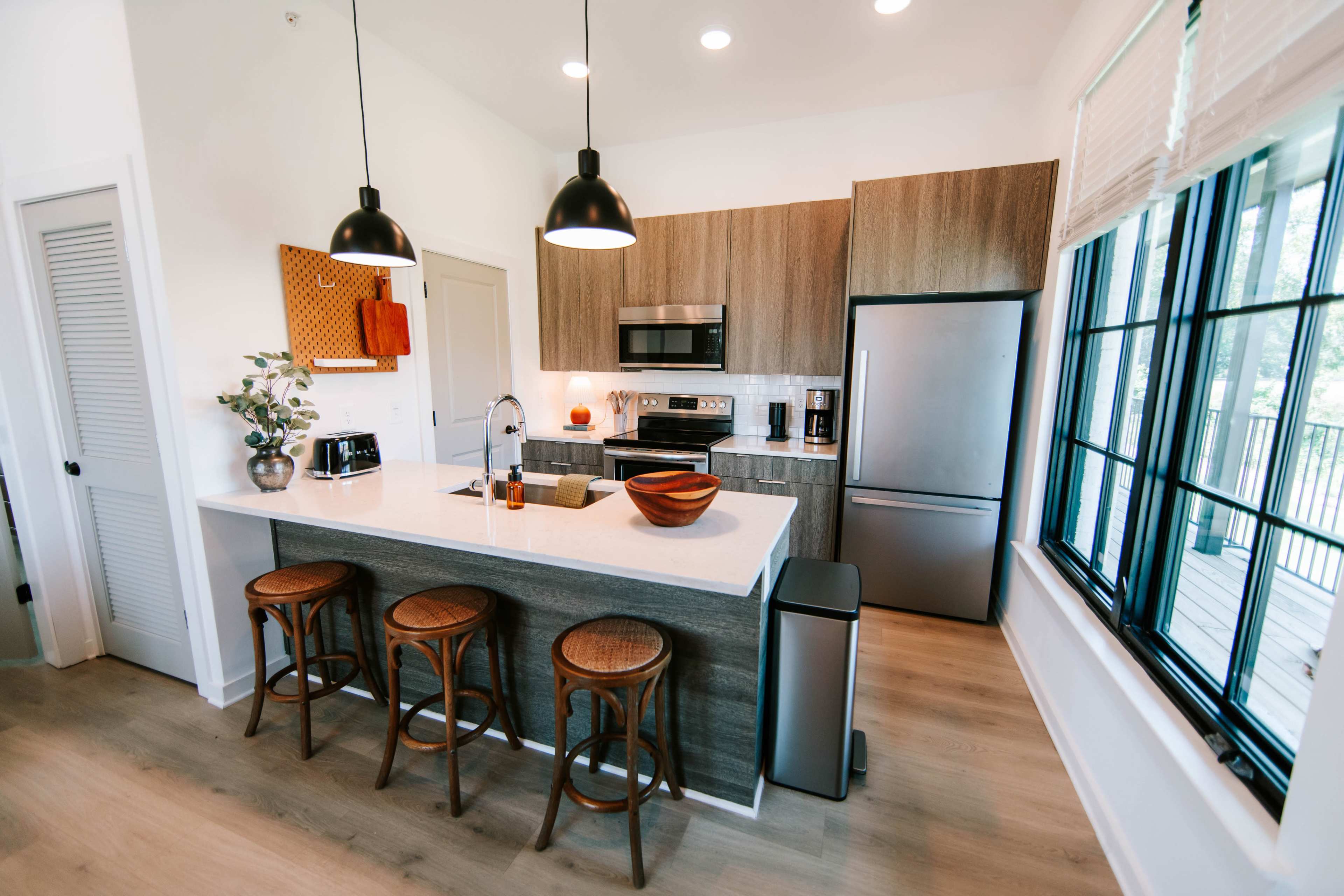 The image shows a modern kitchen with wooden cabinetry, stainless steel appliances, and three bar stools at a counter.
