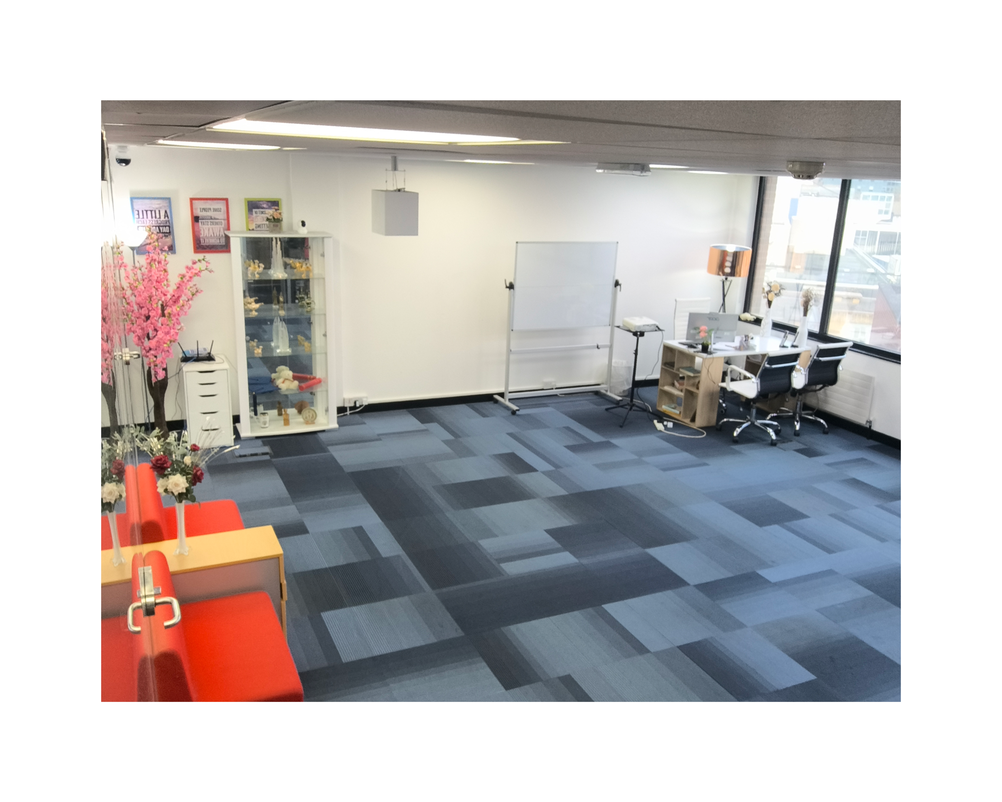 The image shows a spacious office interior with a desk, whiteboard, and display case, featuring blue carpet tiles and large windows.
