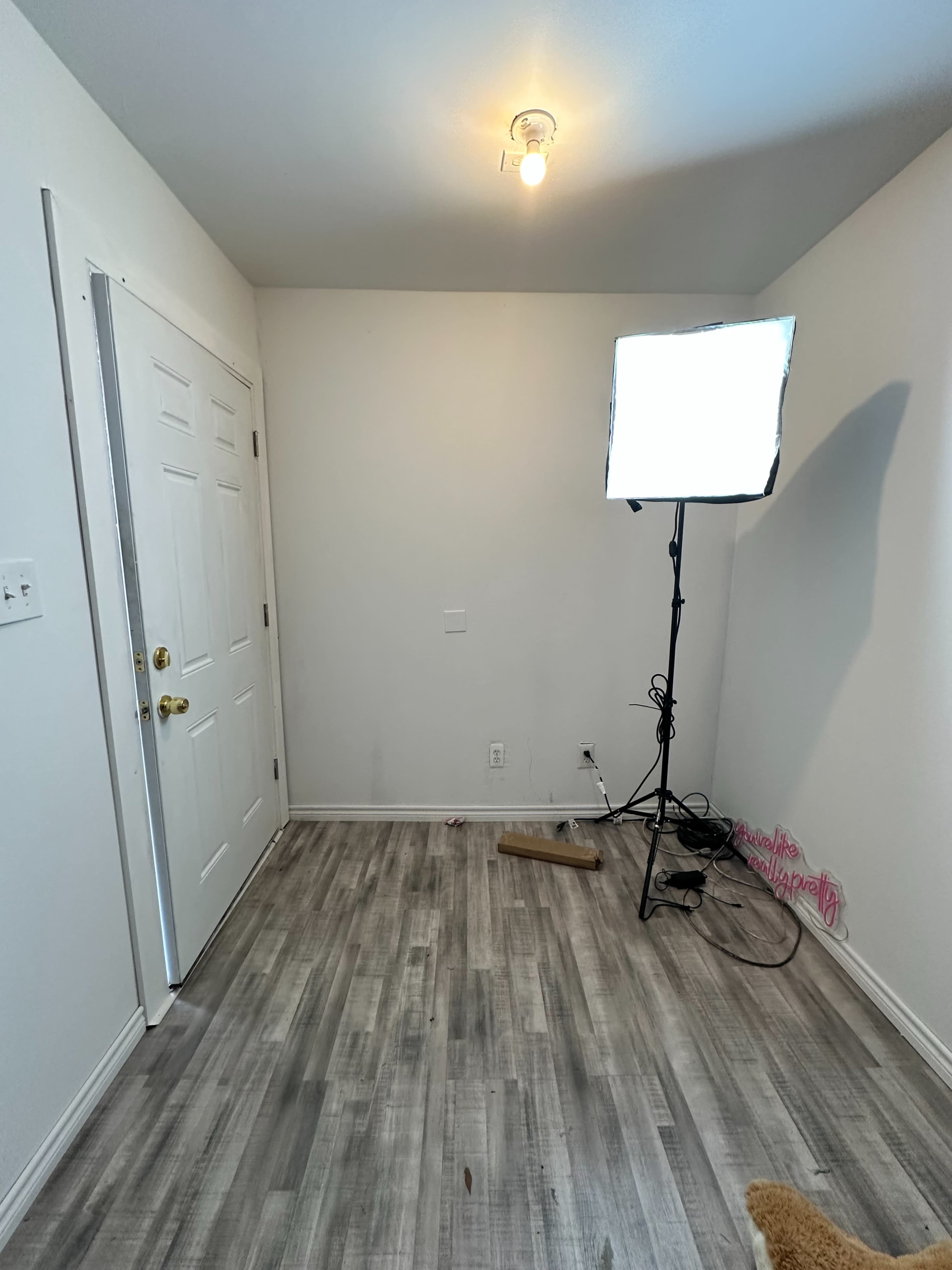 A small, empty room with a door on one wall, a light stand with a softbox, and gray wooden flooring.