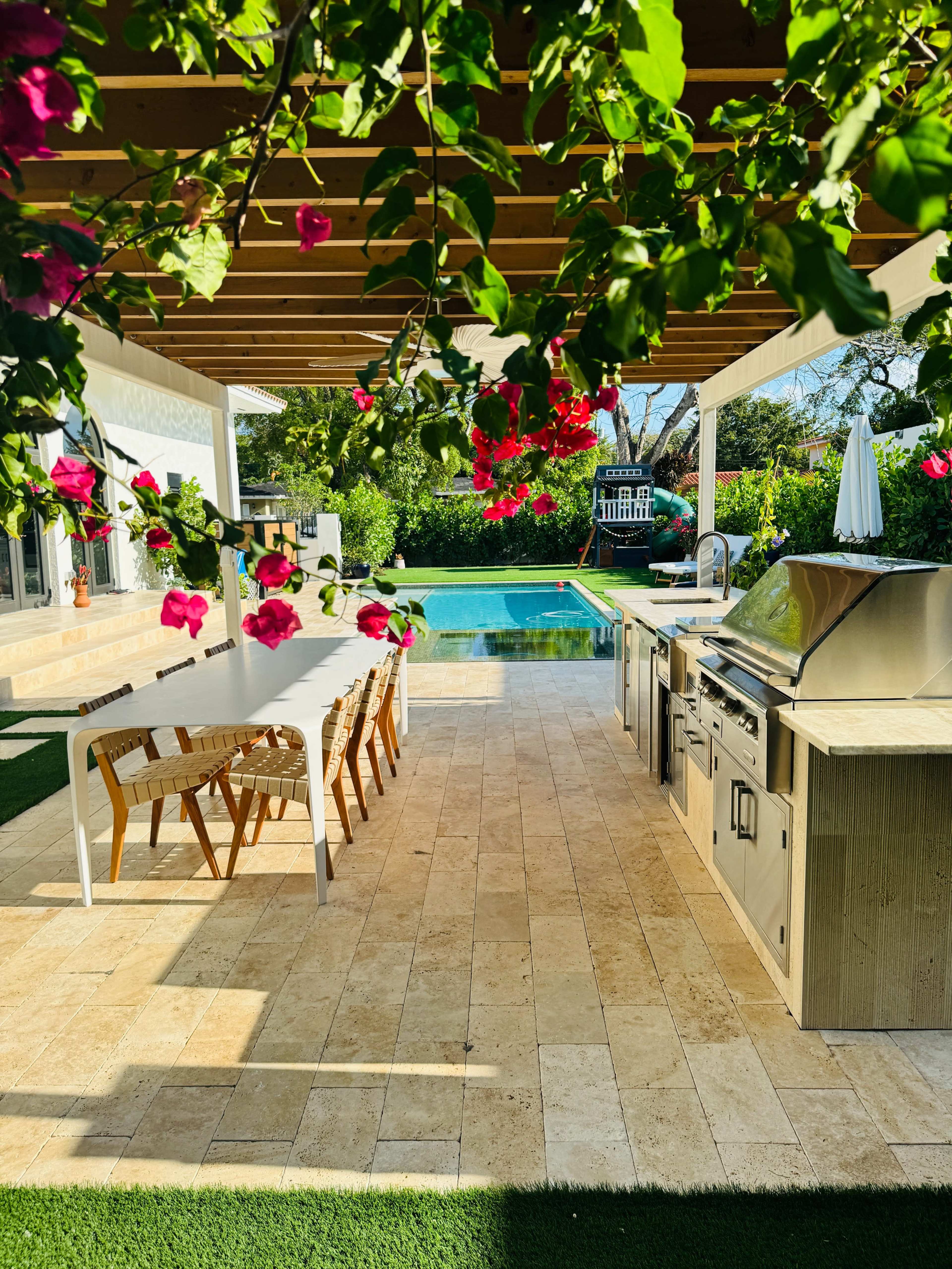 A modern outdoor kitchen and dining area with a view of a swimming pool, surrounded by lush greenery and blooming flowers.