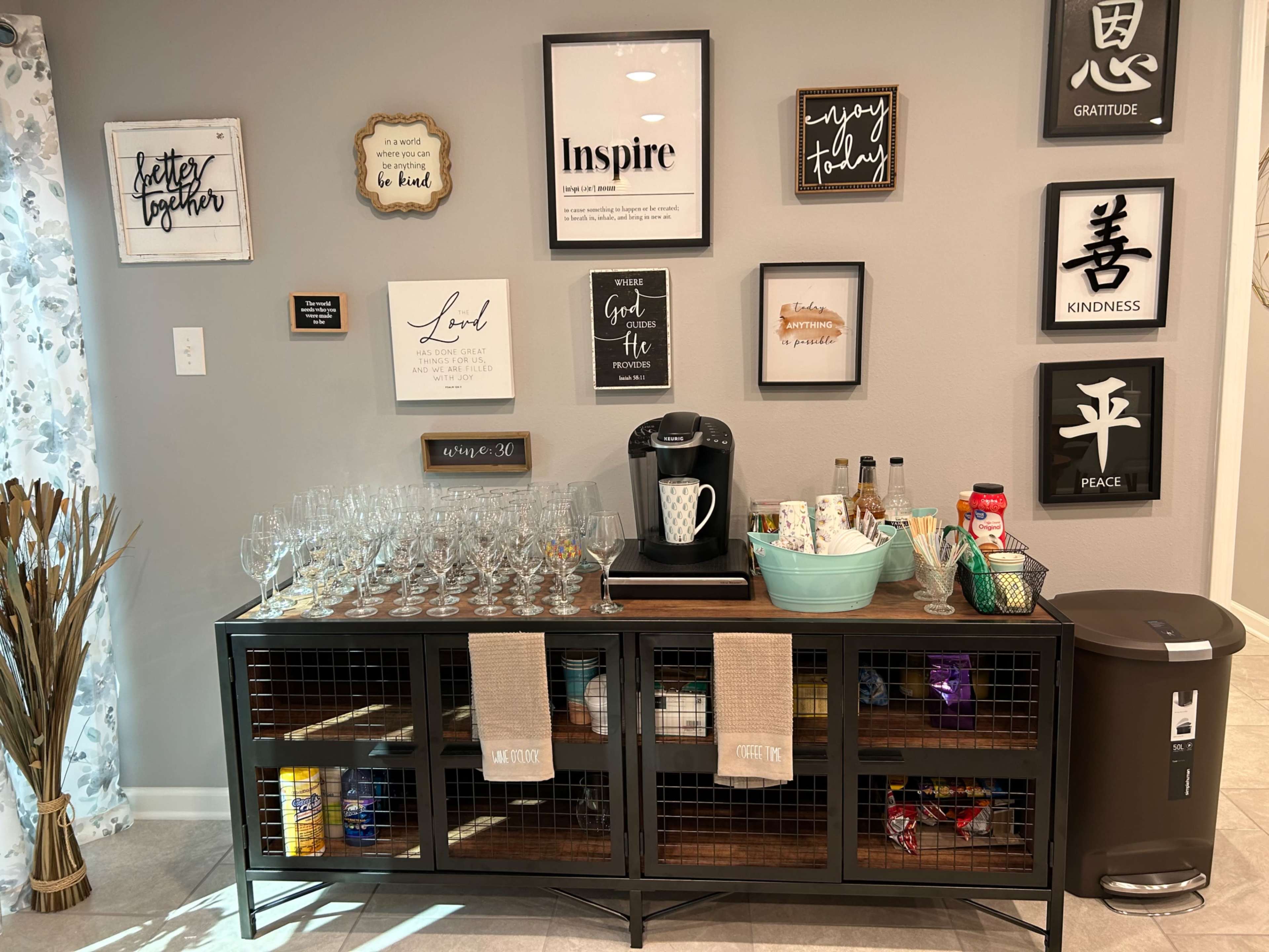 A neatly organized home bar cabinet with glassware, a coffee machine, various beverages, and decorative wall art featuring inspirational messages.