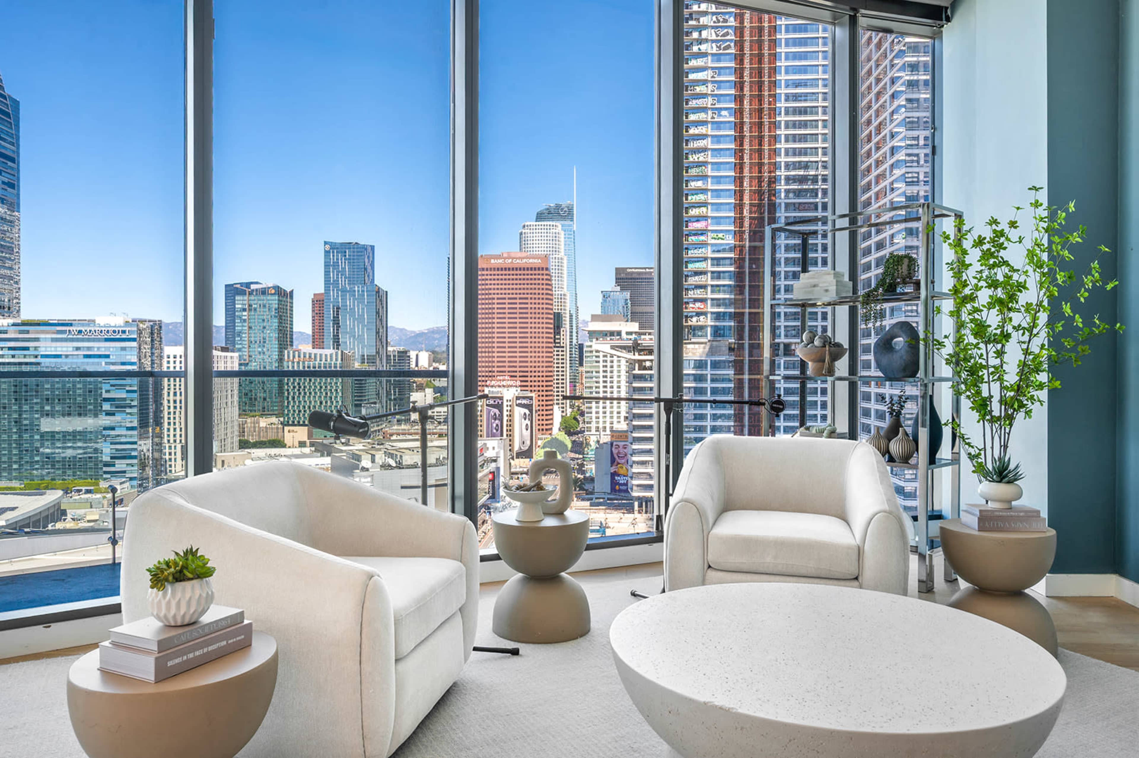 A modern living room features two white armchairs and a round coffee table, with large windows showcasing a skyline view of a city.