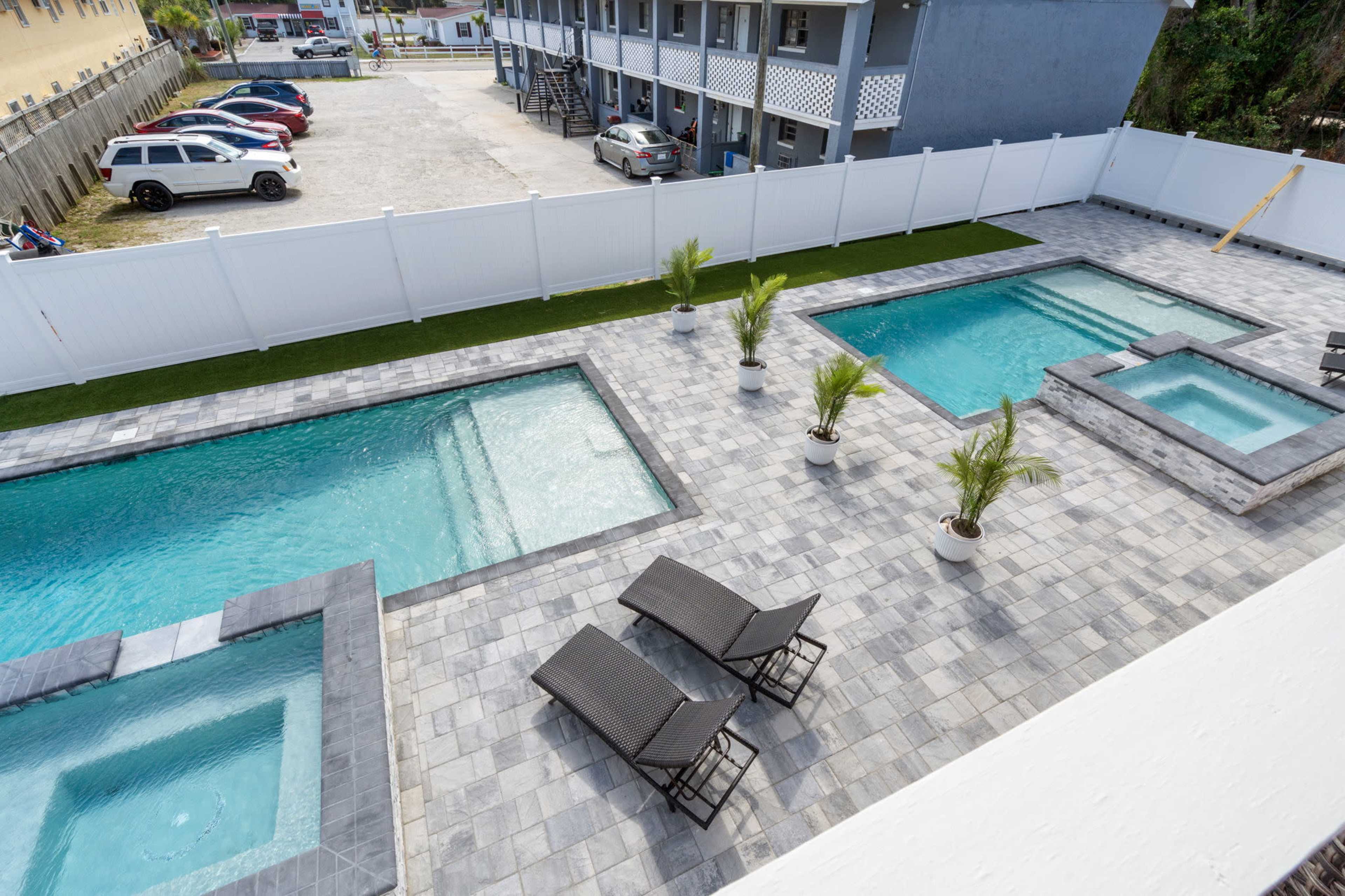 The image shows a fenced pool area featuring multiple rectangular swimming pools, lounge chairs, and potted plants, adjacent to a parking area with parked vehicles.