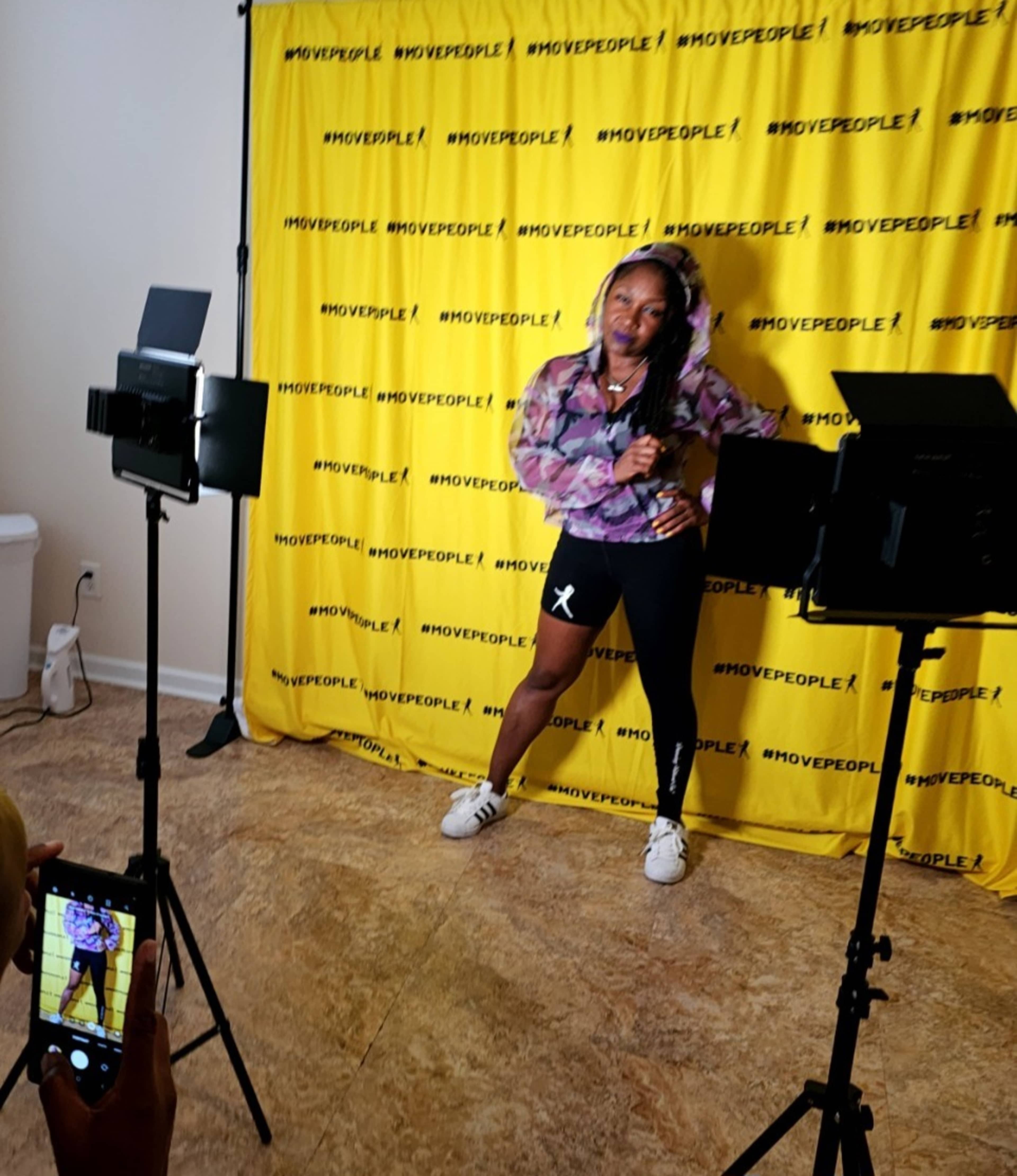 A person poses against a yellow backdrop with the text "#MOVEPEOPLE!" while two mounted lights illuminate the scene.
