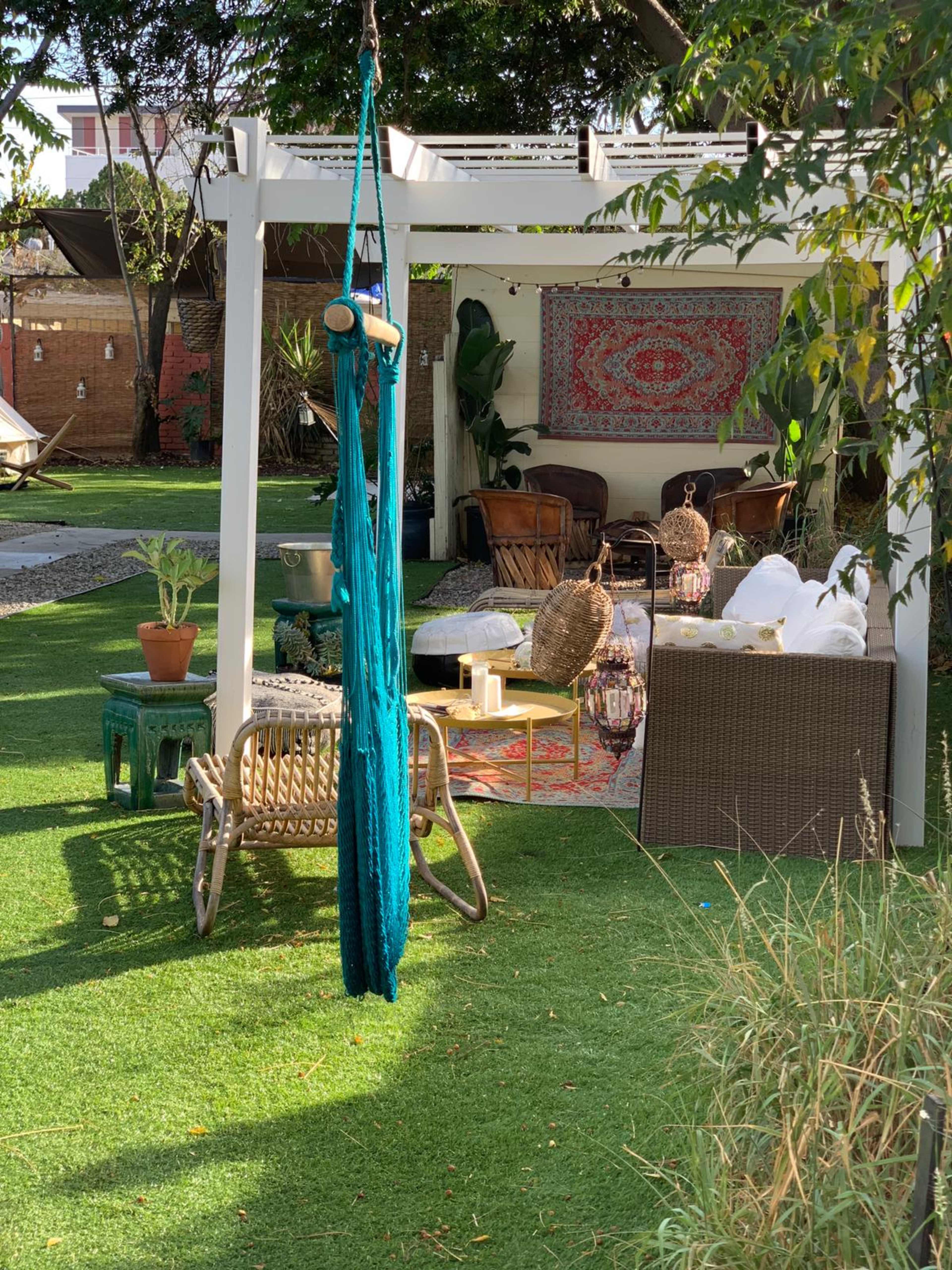 A green lawn with a hammock hanging from a white structure, surrounded by various styled seating areas and decorative elements under a bright sun.