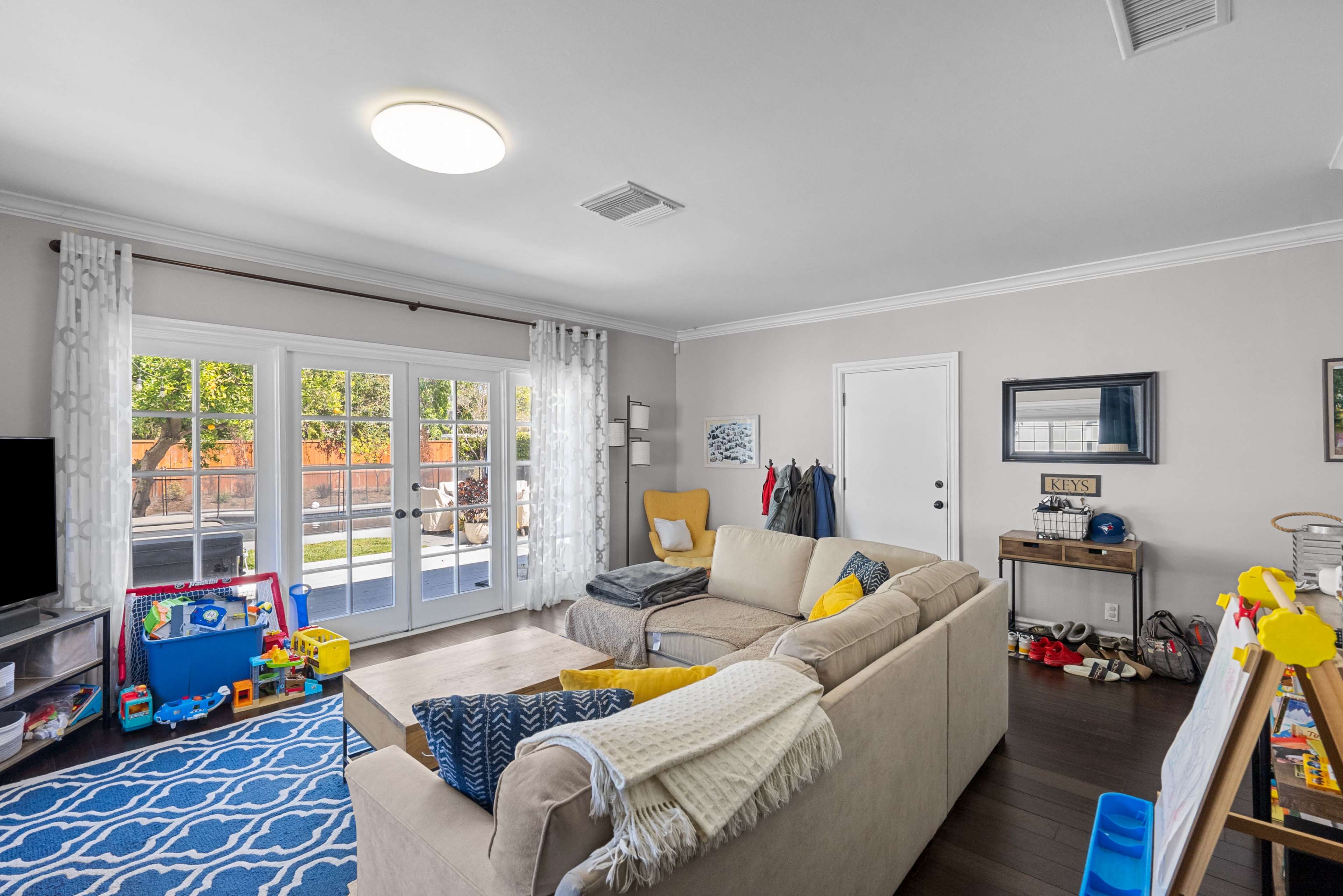 A cozy living room features a beige sectional sofa, a patterned blue rug, a TV stand with toys, and double glass doors leading to an outdoor area.