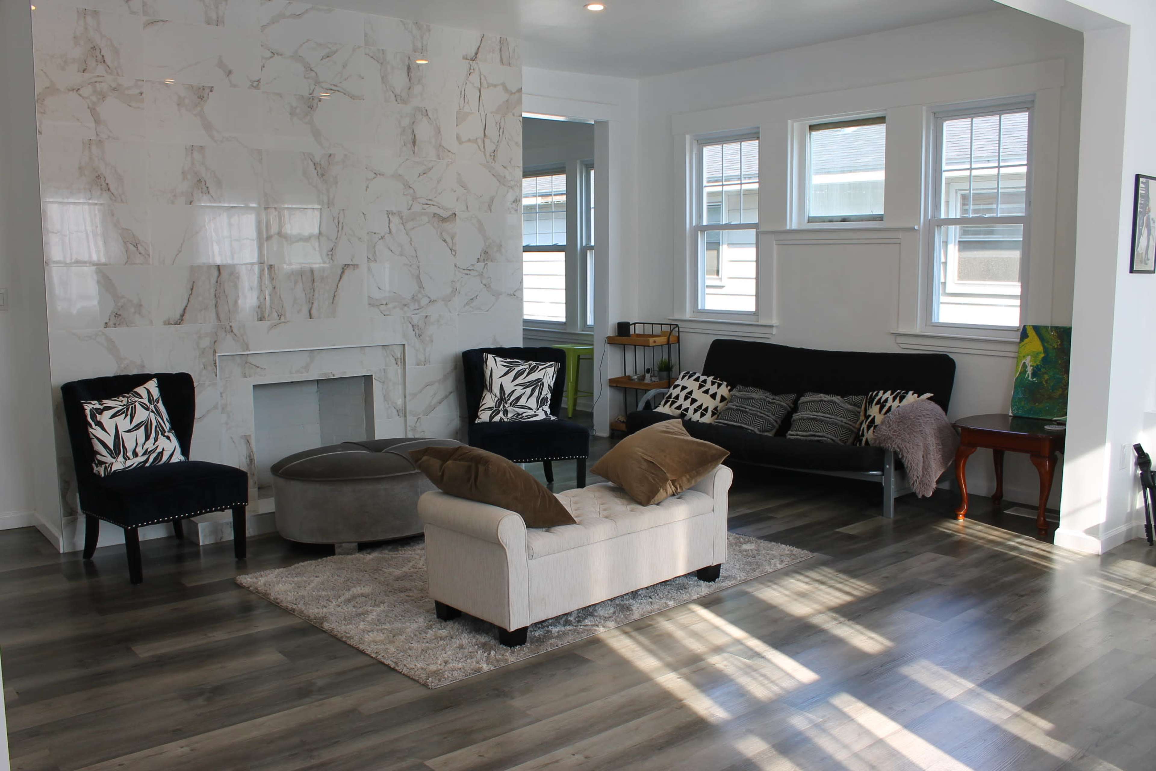 The room features modern furniture, including a black futon, two patterned chairs, and a light-colored couch, arranged around a rug on a floor with gray planks, alongside a marble fireplace.