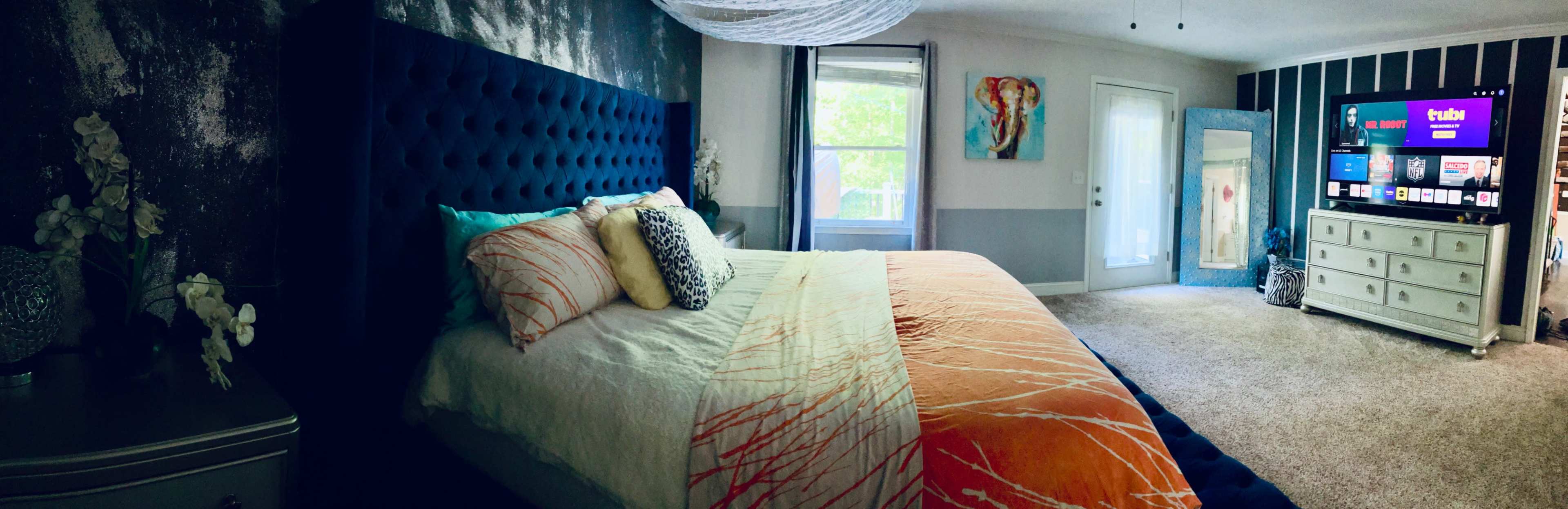 The image shows a spacious bedroom featuring a large tufted bed with an orange and white bedspread, a flat-screen TV mounted on the wall, and a mirrored dresser.
