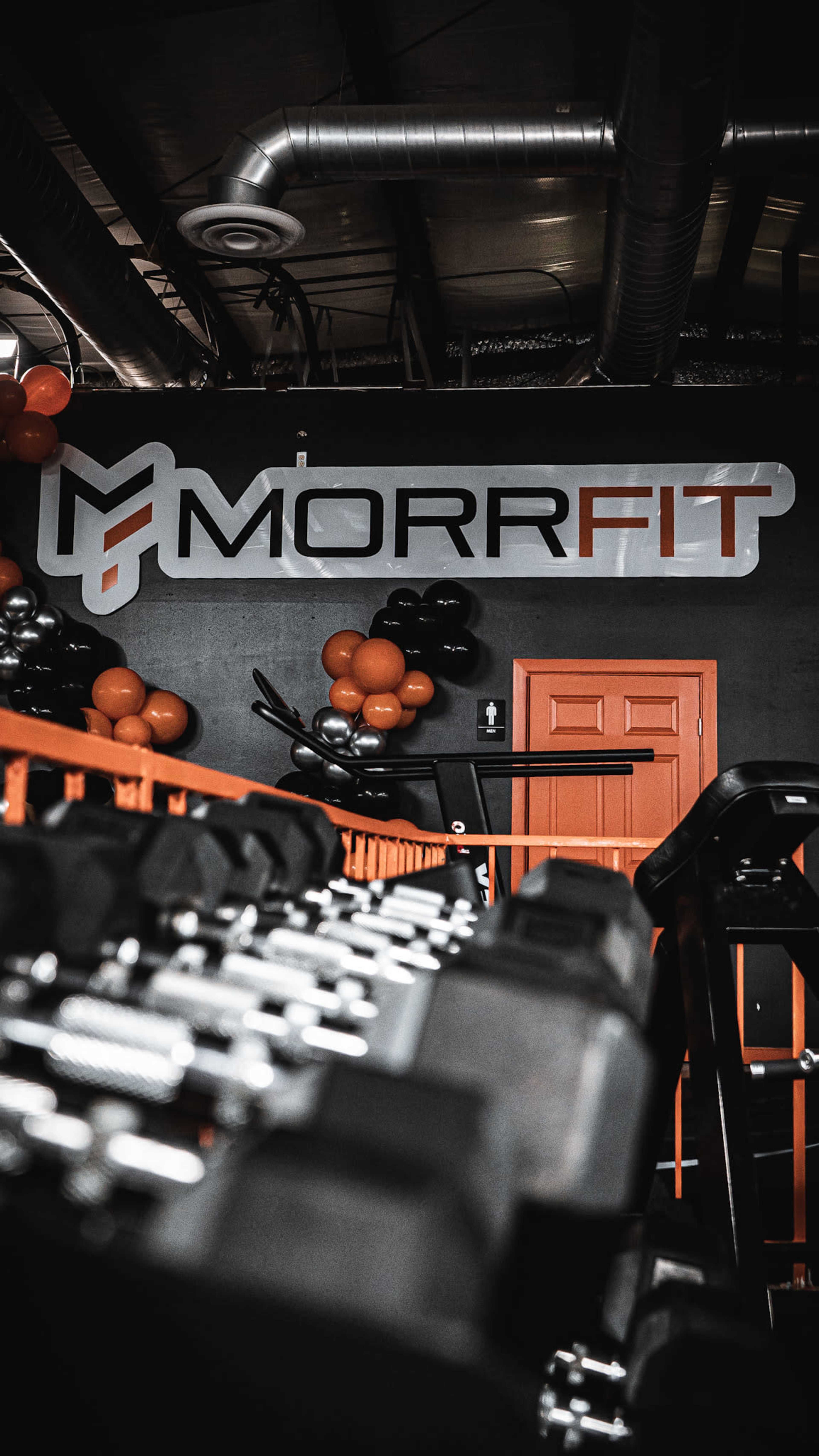 A gym interior featuring a wall with the "MORRFIT" logo and a rack of dumbbells in the foreground.