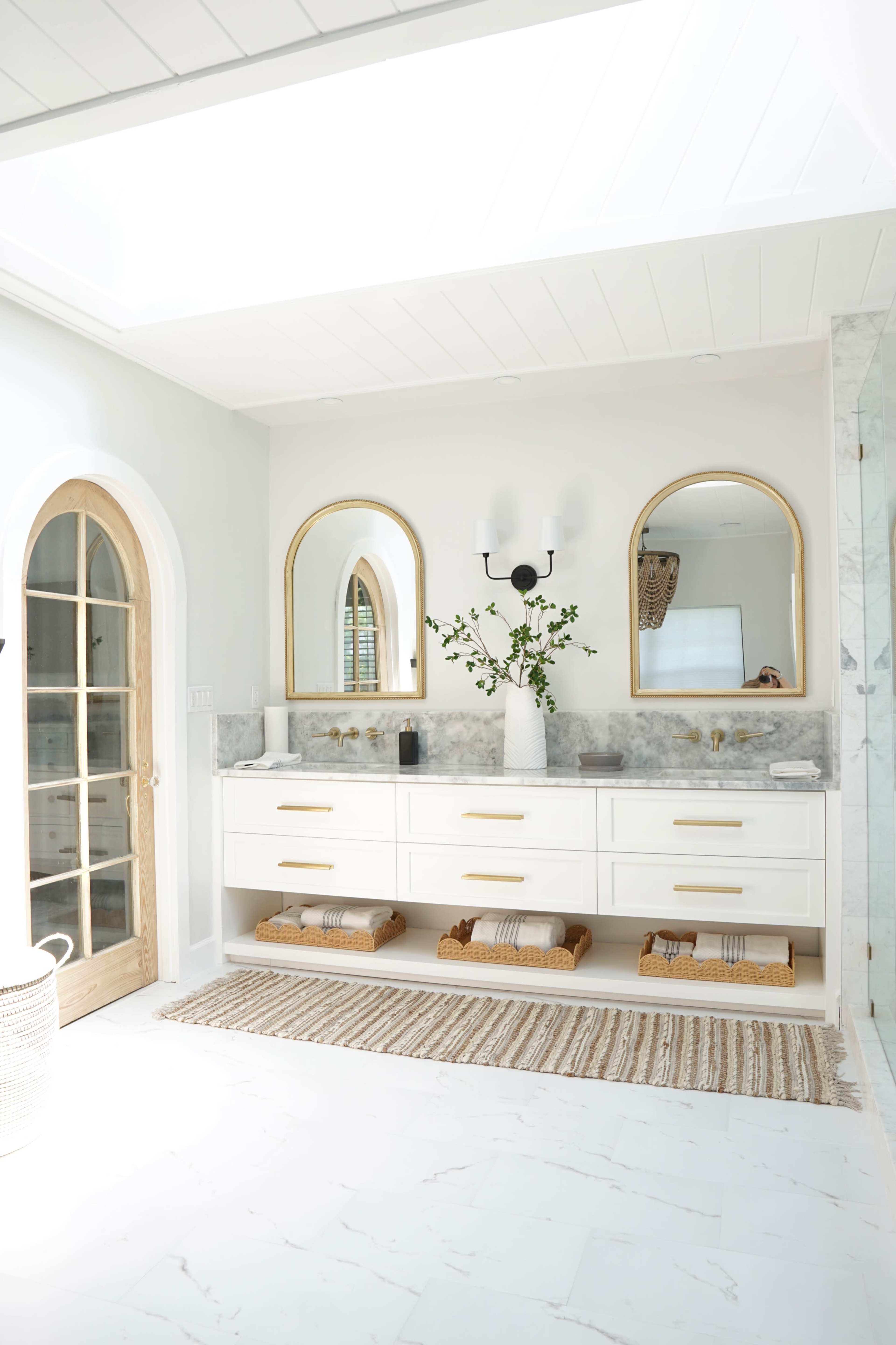A modern bathroom features a double sink vanity with marble countertops, three mirrors above, and a large window providing natural light.