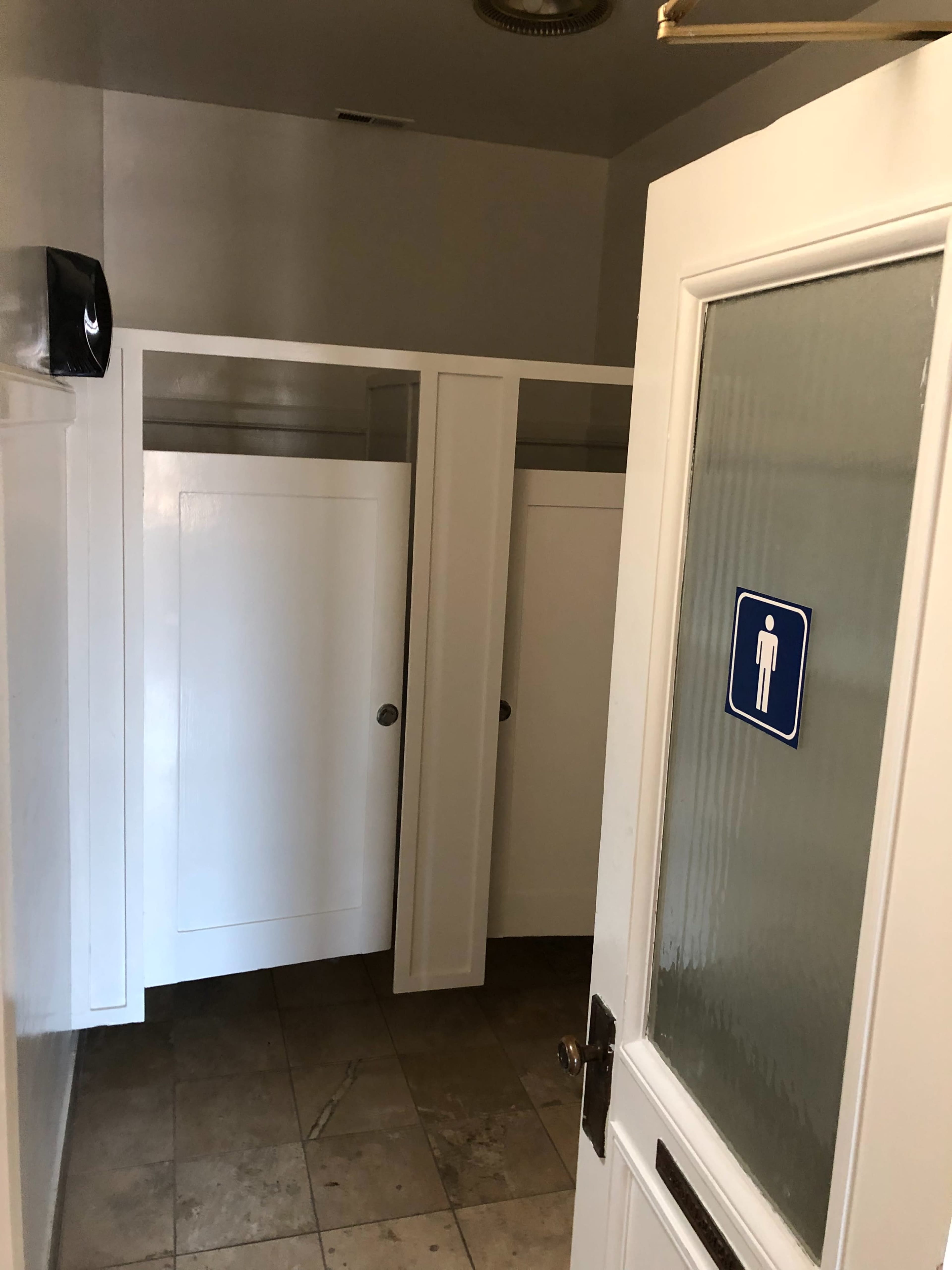 The image shows an interior view of a restroom with several white stalls and an open door leading outside.