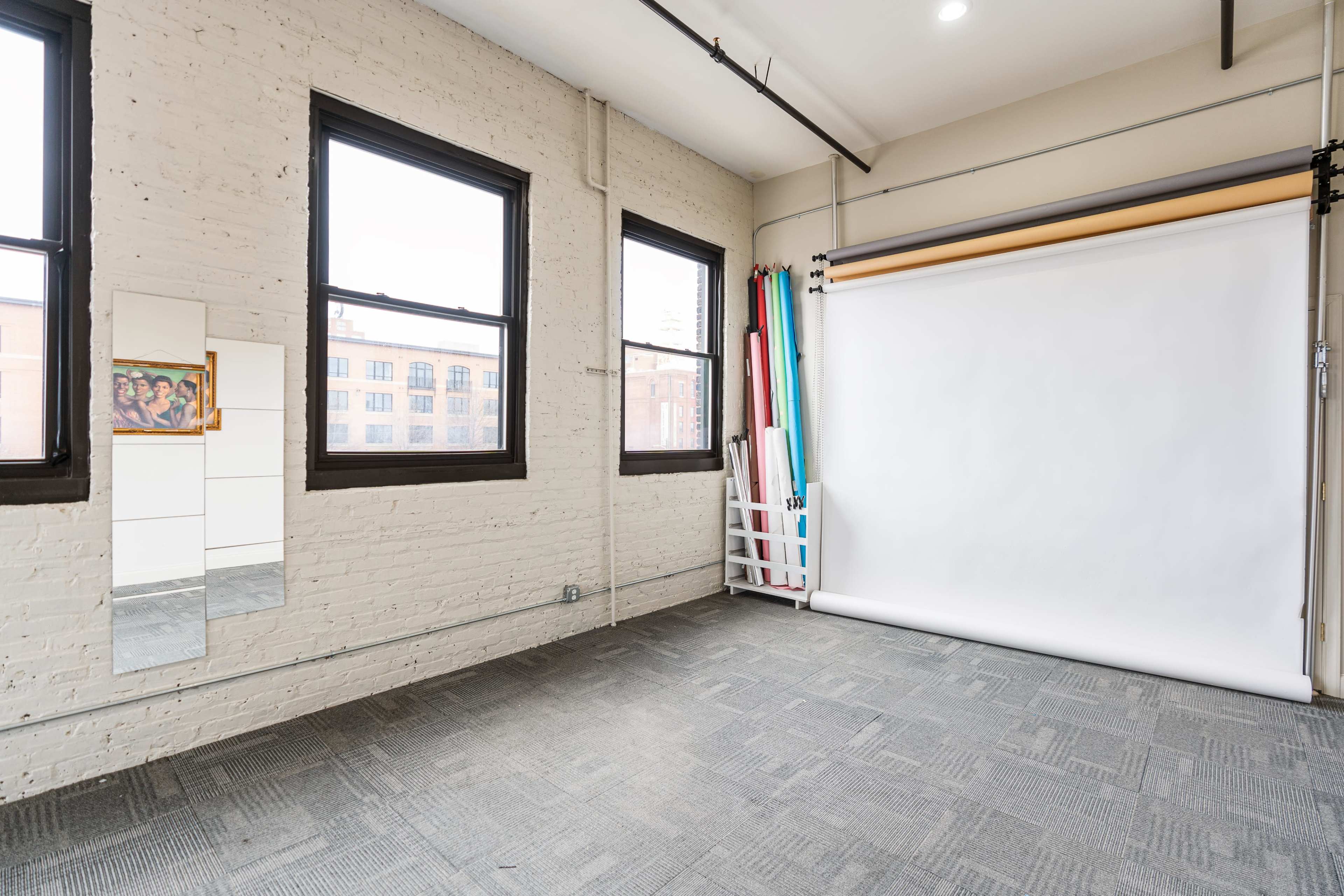 A bright studio space with a white backdrop, large windows, and a mirror on the wall.