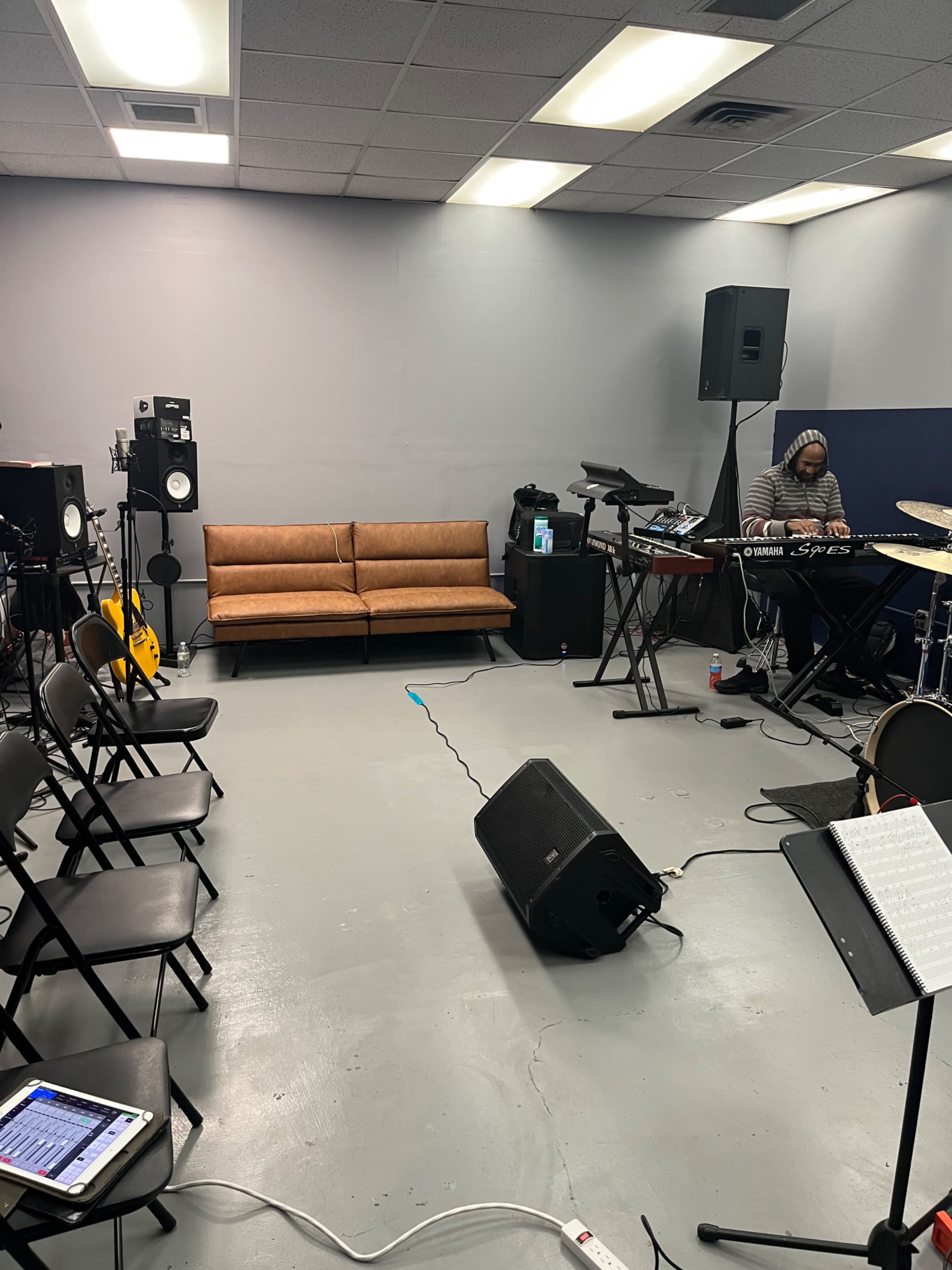 The image shows a rehearsal space featuring a brown couch, musical instruments, a sound system, and several black folding chairs arranged in front of a keyboardist.