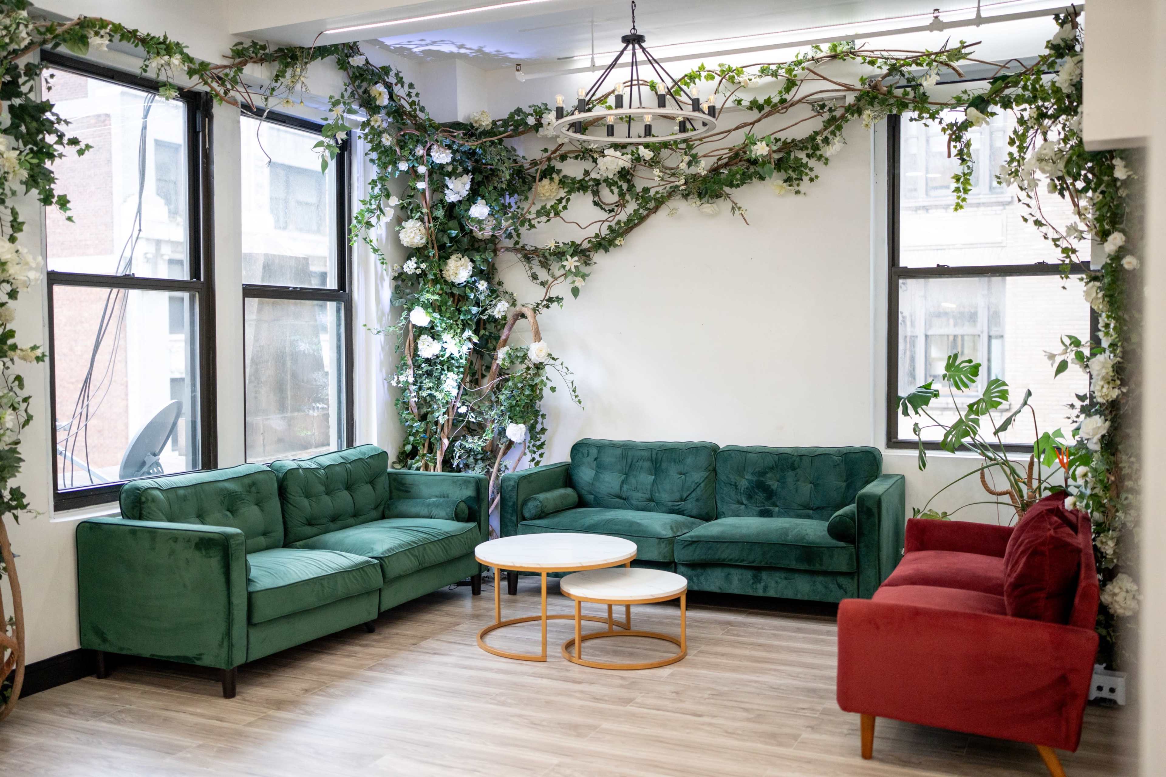 The image shows a cozy lounge area featuring green sofas, a red sofa, a circular table, and decorative plants along the walls.