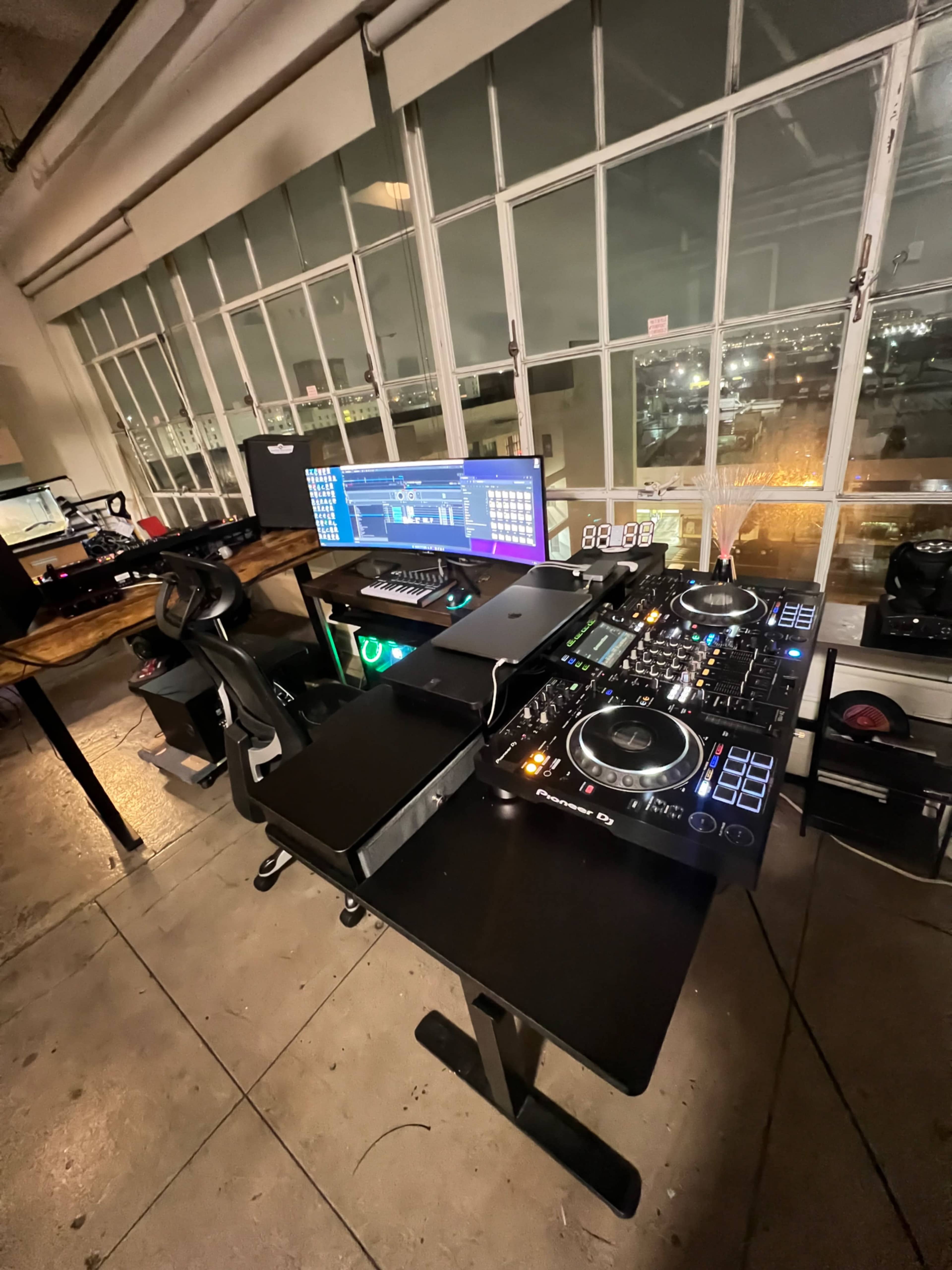A DJ setup with a controller, dual monitors, and a laptop is positioned in a well-lit room featuring large windows overlooking a cityscape at night.