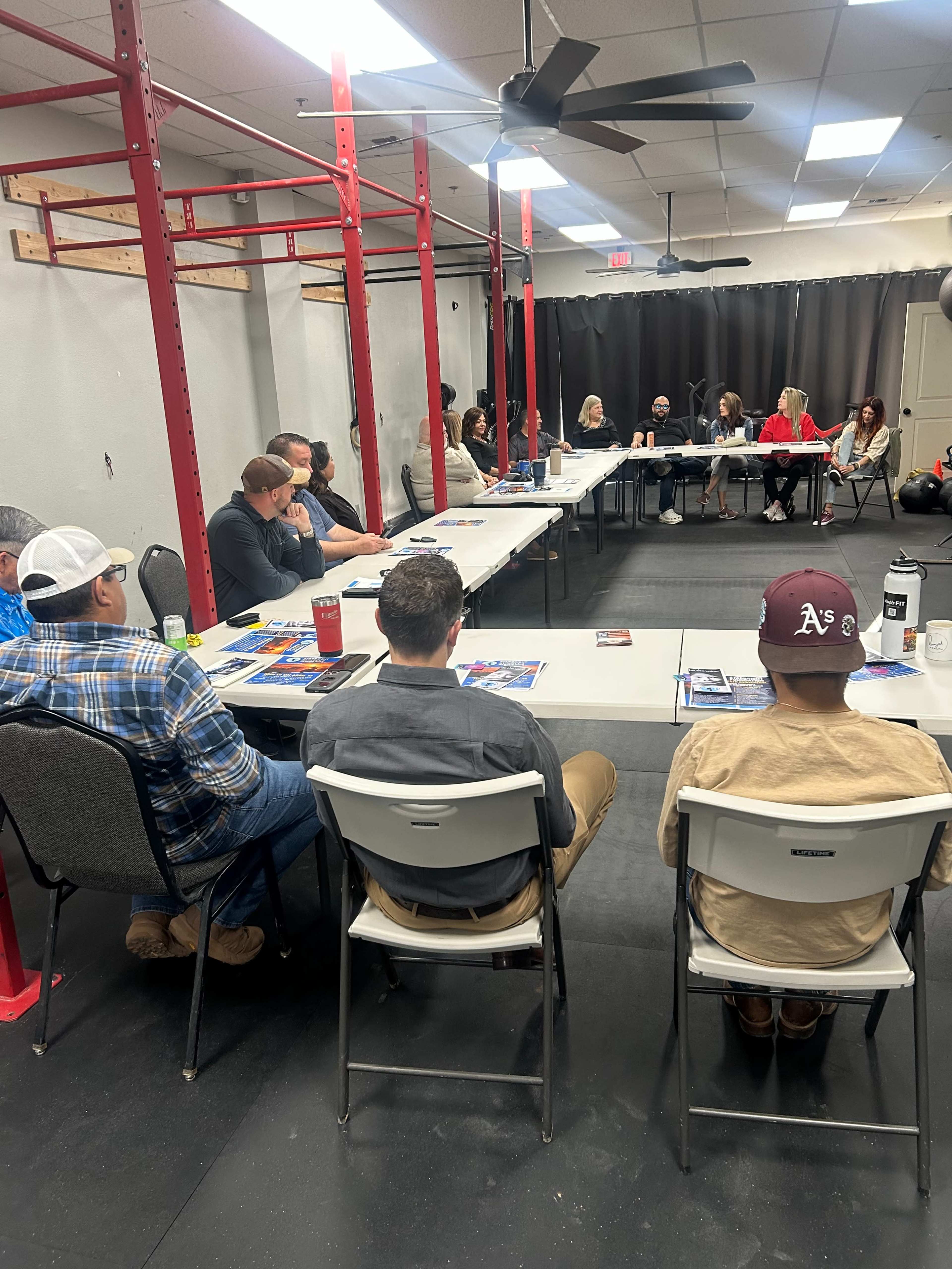 A group of people is seated around tables in a room set up for a meeting, with red exercise equipment in the background.