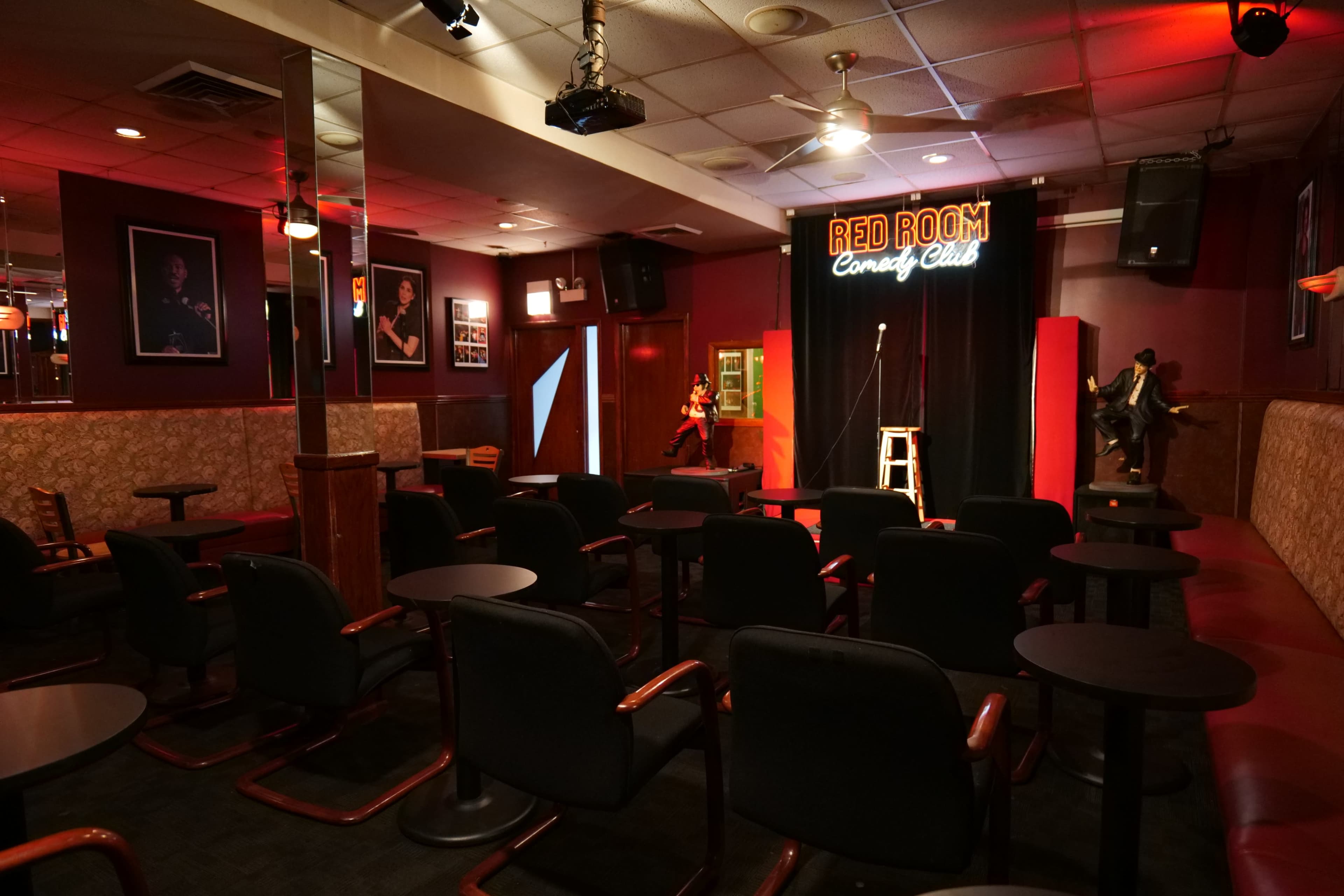 A comedy club with empty chairs arranged in front of a stage featuring a microphone and a stool, illuminated by a sign that reads "RED ROOM Comedy Club."