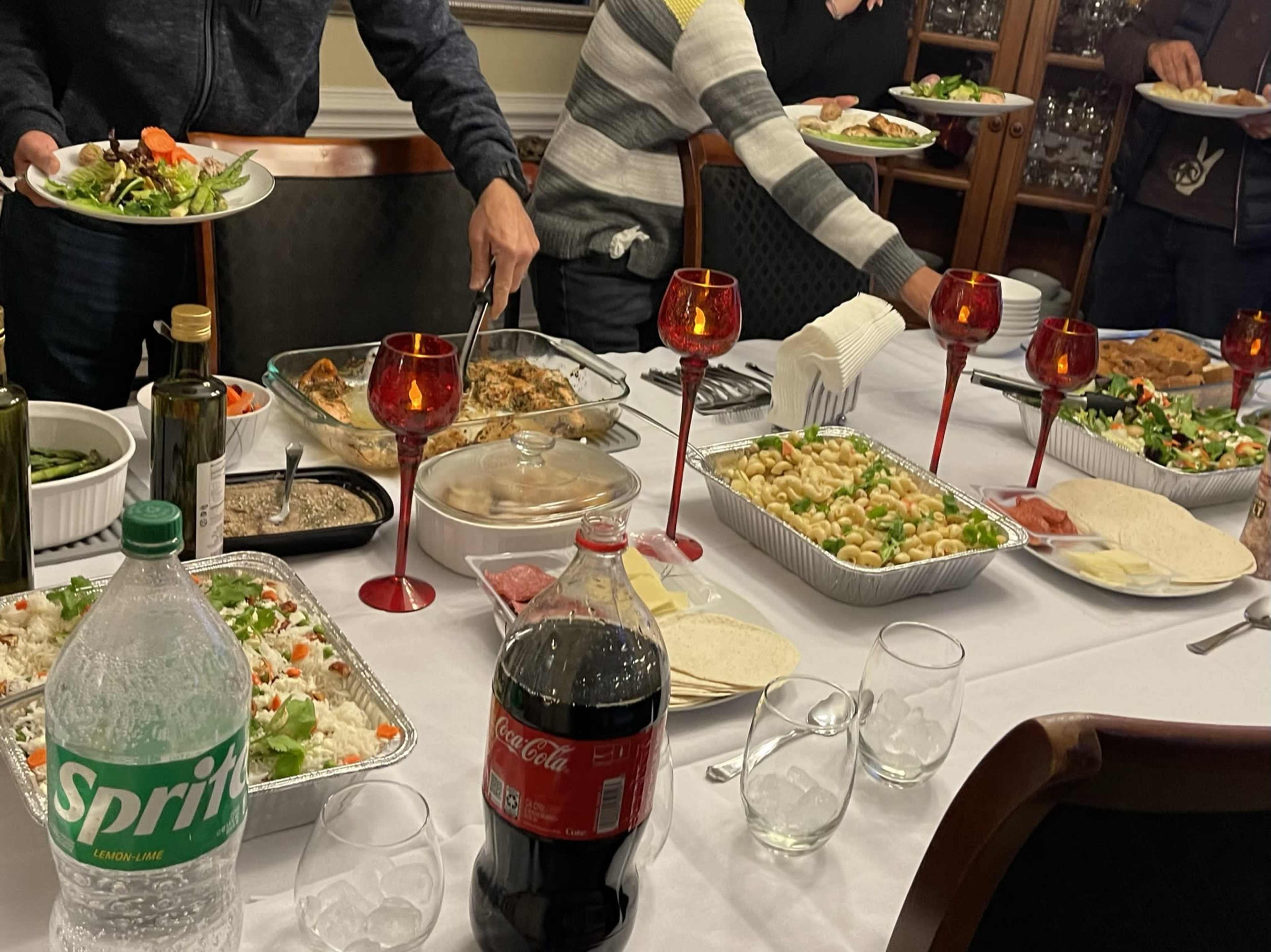 A dining table is set with various food dishes, drinks, and people serving themselves.