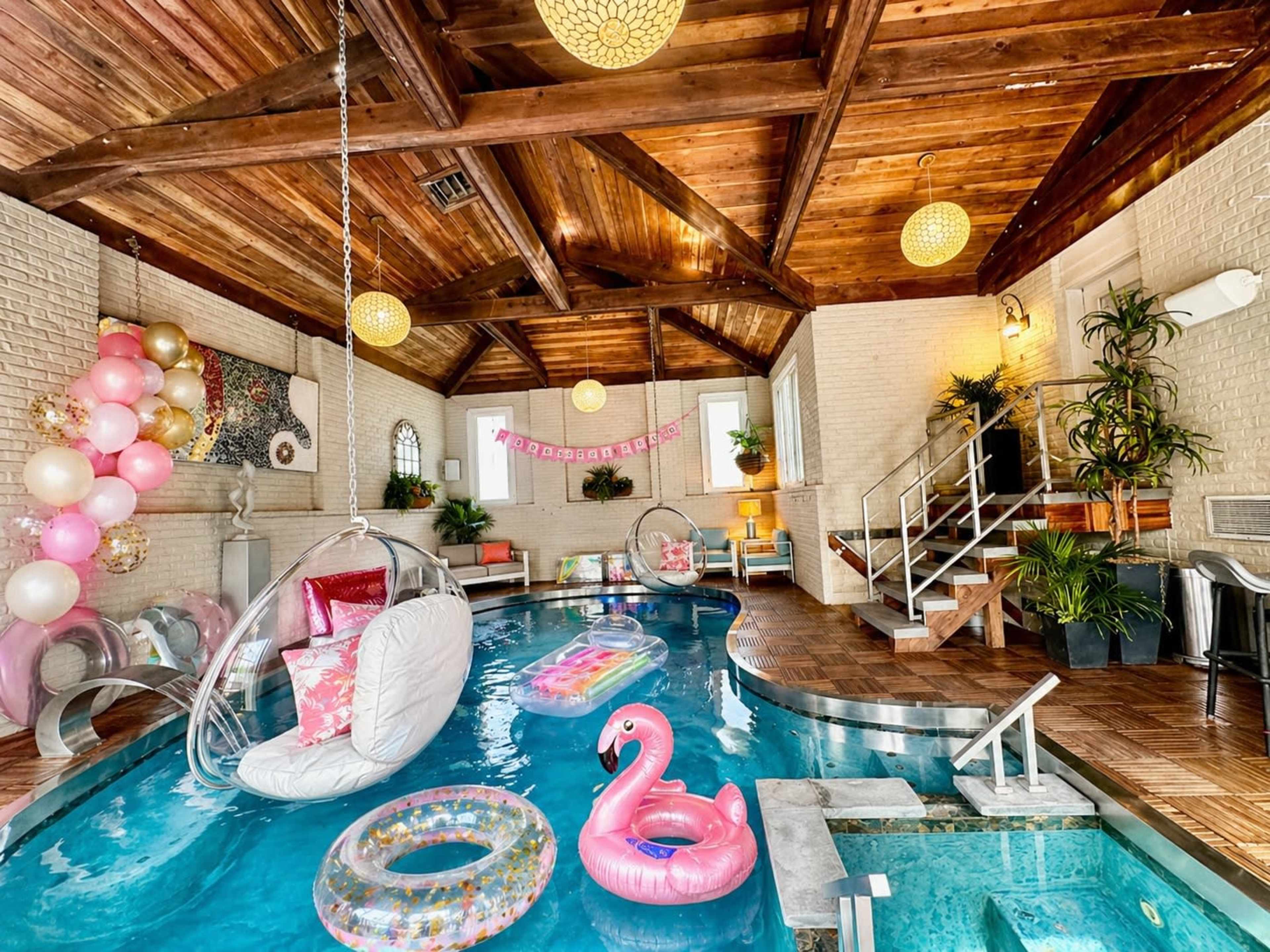 The image features a modern indoor pool area with a wooden ceiling, colorful floating pool toys, and decorative elements including balloons and plants.