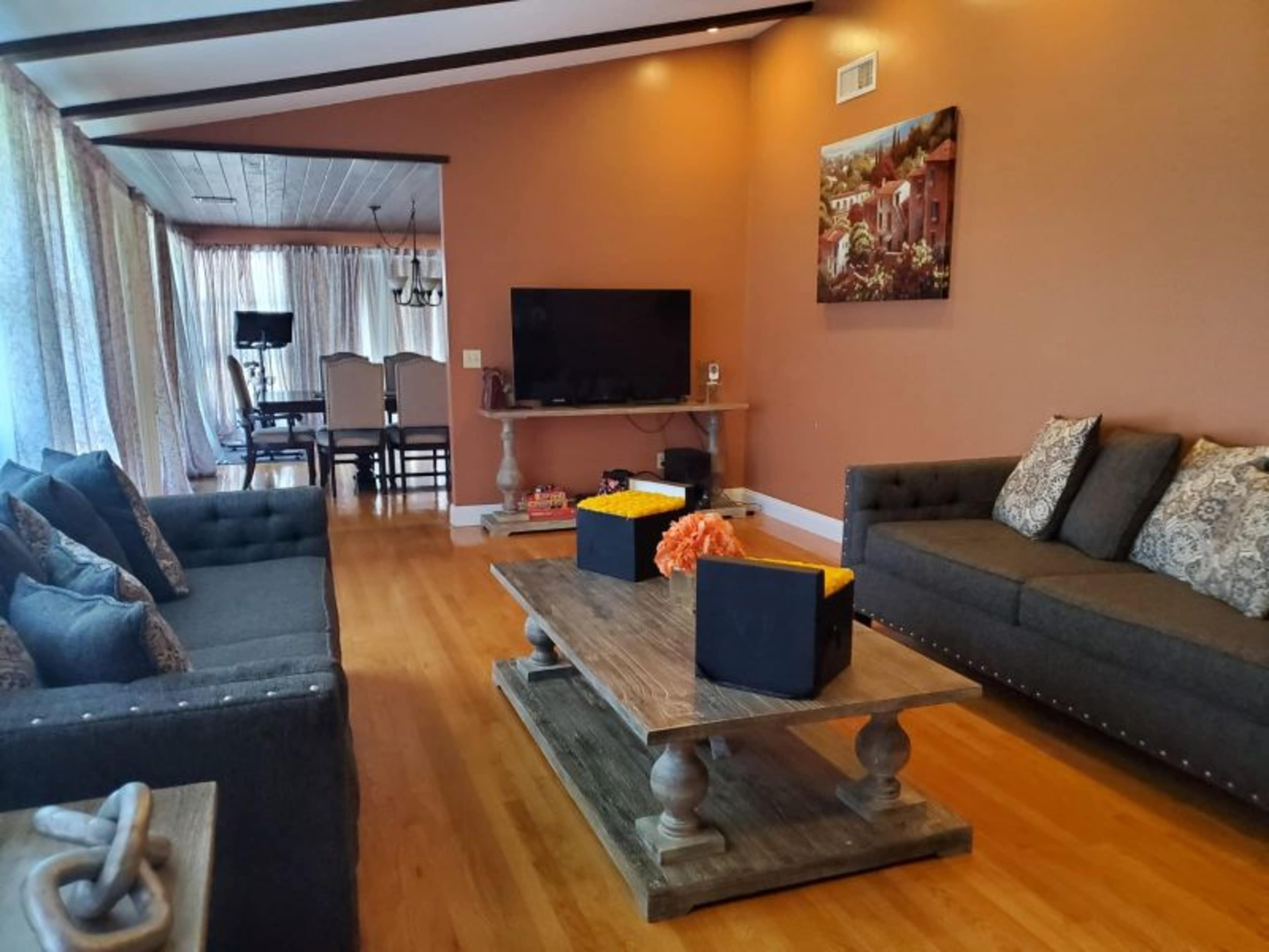 A living room with two sofas, a wooden coffee table, and a television, surrounded by large windows with sheer curtains and a view of a dining area.