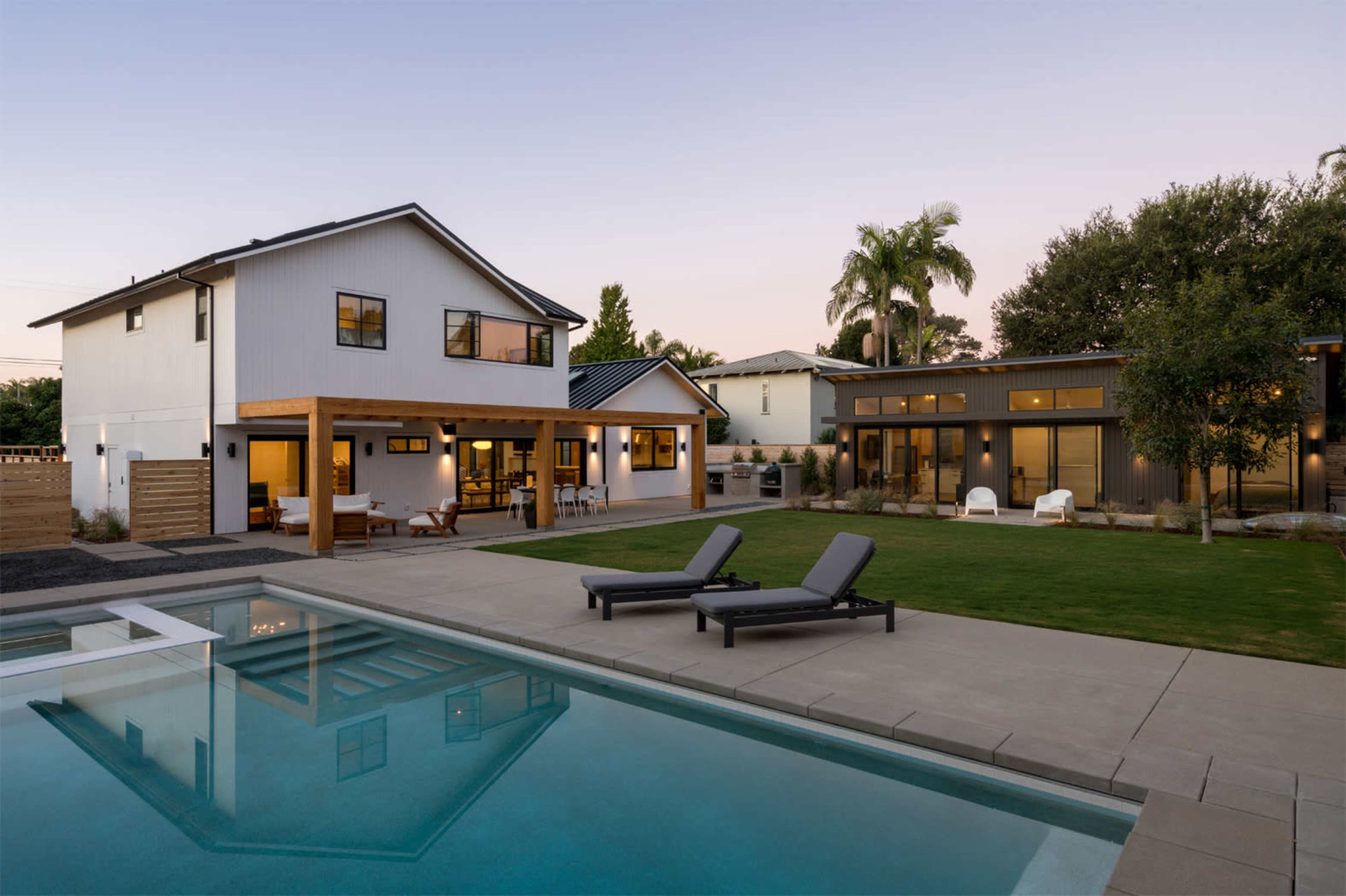 A modern backyard features a pool with lounge chairs, adjacent to two contemporary-style houses surrounded by greenery at dusk.